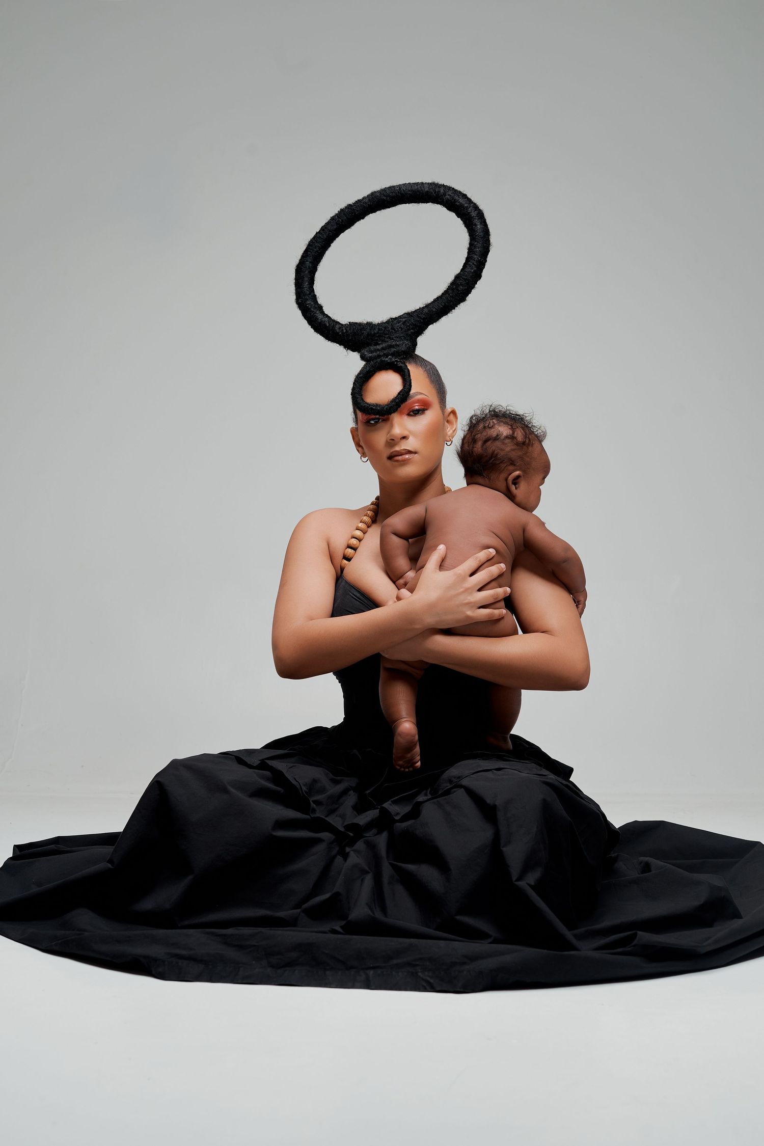 Woman in black dress with avant-garde halo headpiece holds a baby in a dramatic studio portrait.