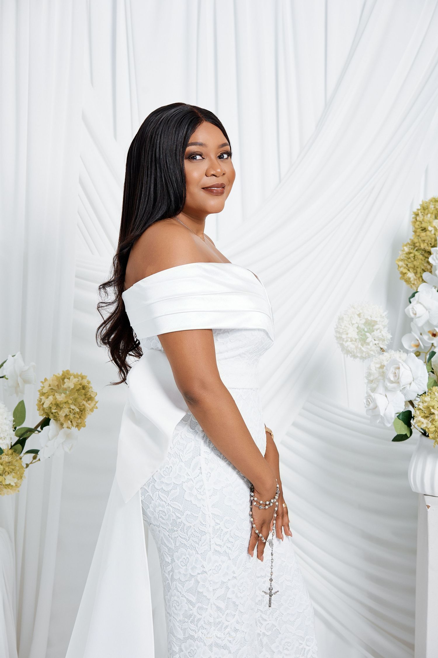 Elegant Black woman in off-shoulder white mermaid gown posing beside white floral arrangements on draped backdrop.