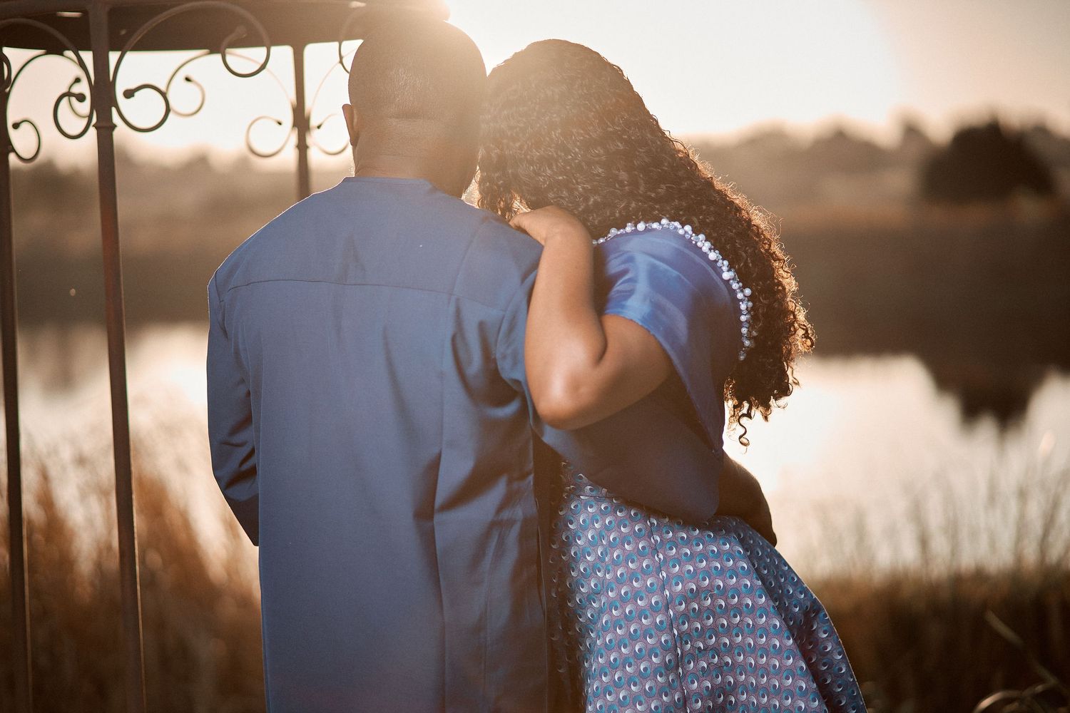Romantic couple sharing an intimate moment by a sunlit lake at golden hour, viewed from behind.