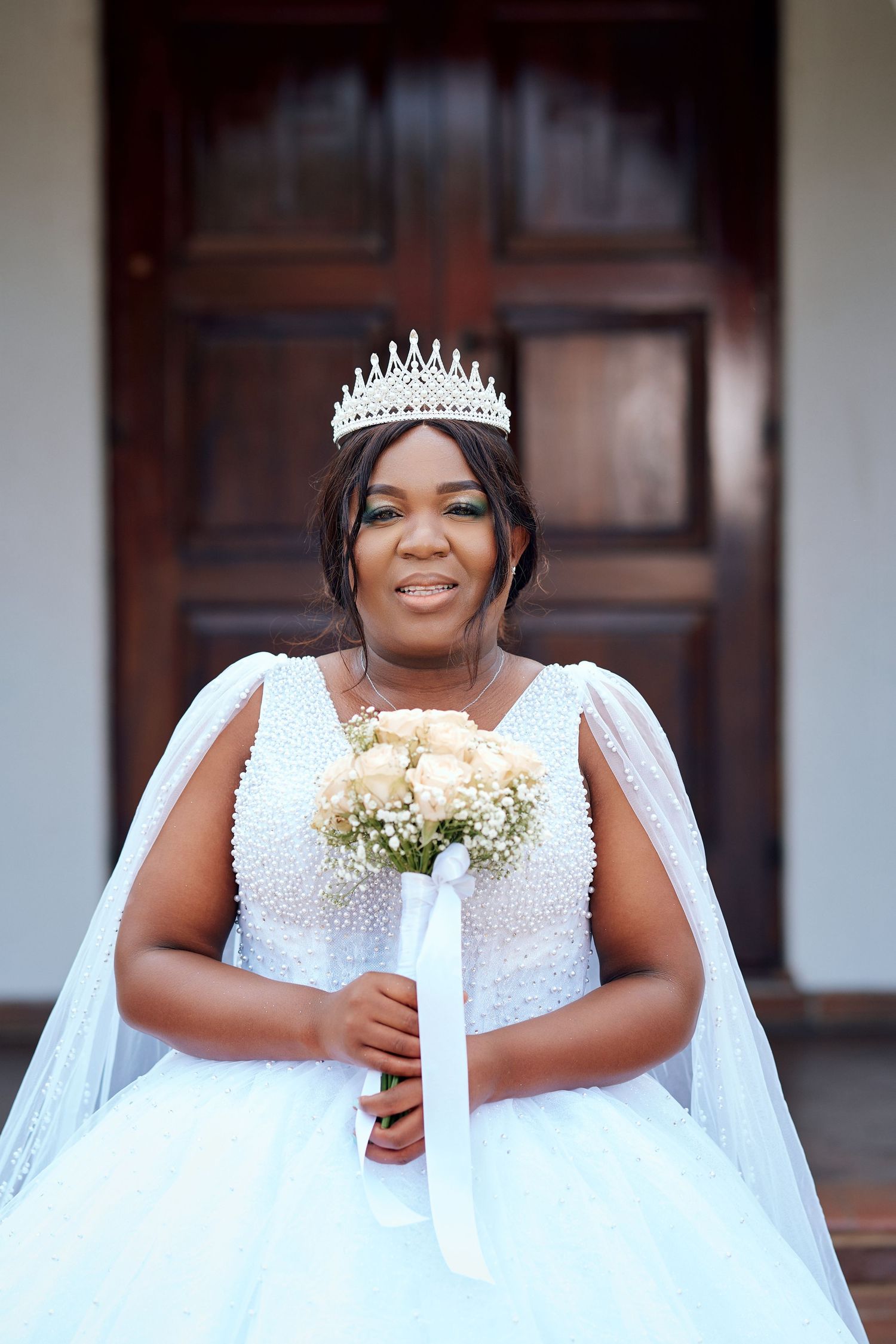 Beautiful Black bride smiling in white pearl wedding gown, crystal crown, holding rose bouquet outdoors.