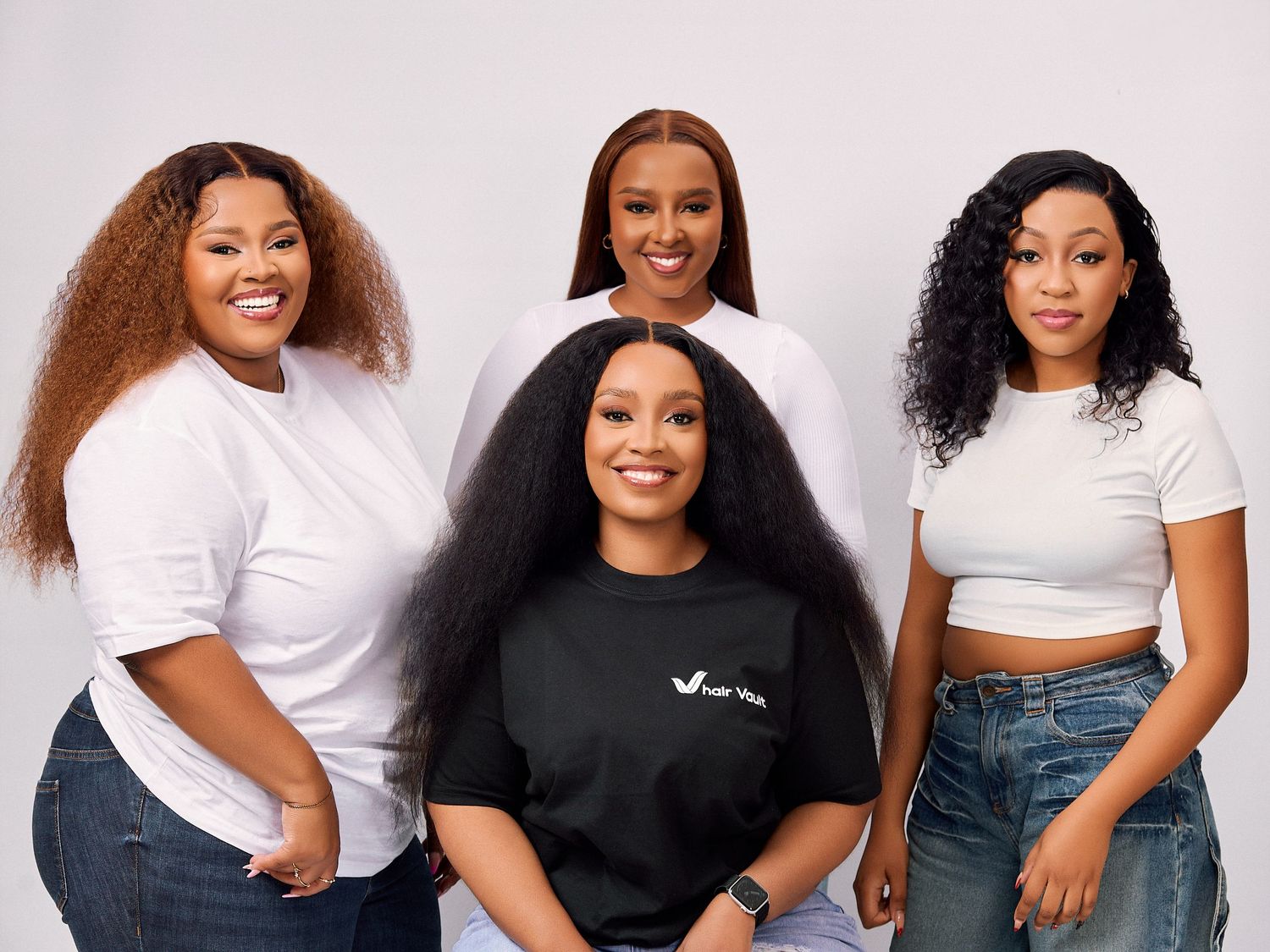 Four Black women in white tops and jeans smiling together in a professional studio portrait photo.