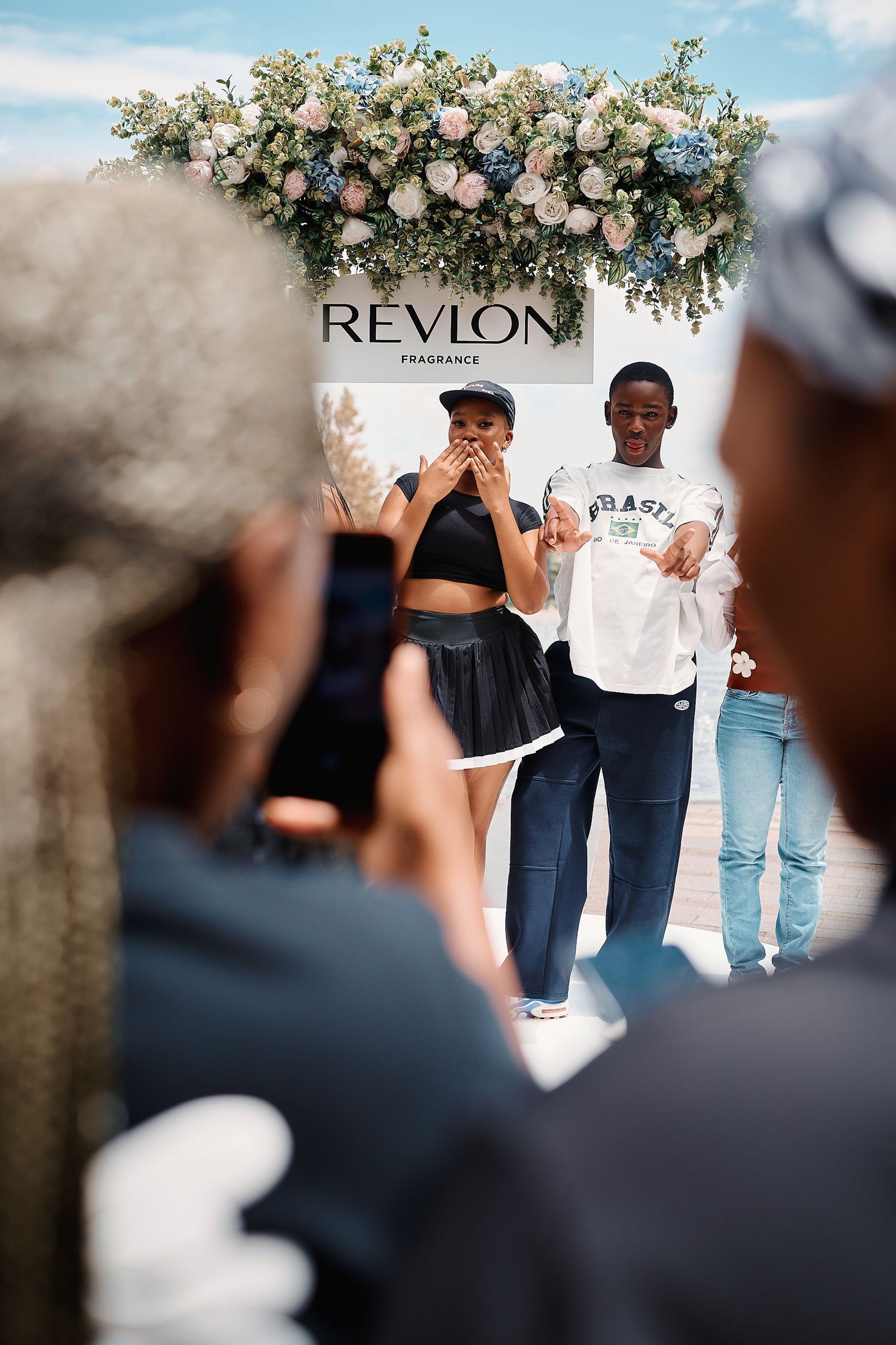 Two people stand before Revlon Fragrance floral arch while photographer captures moment at outdoor event.