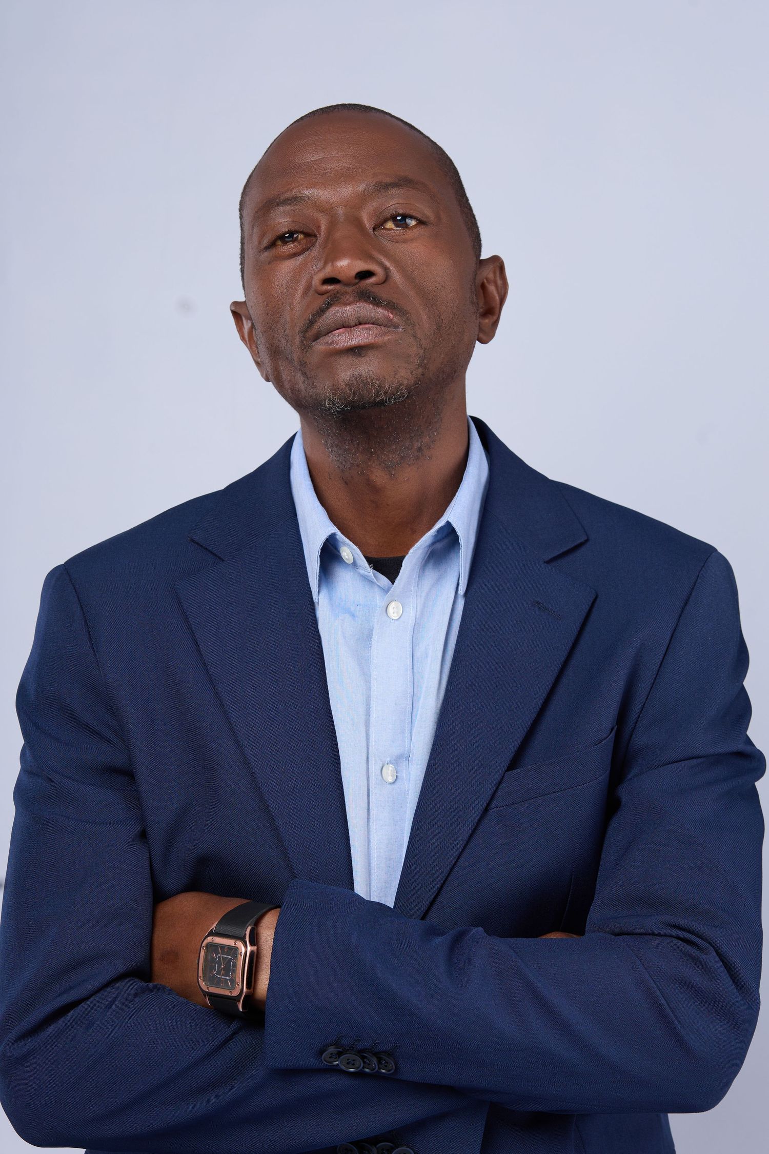 A confident Black man in a navy blue blazer and light blue shirt stands with arms crossed against a white background.