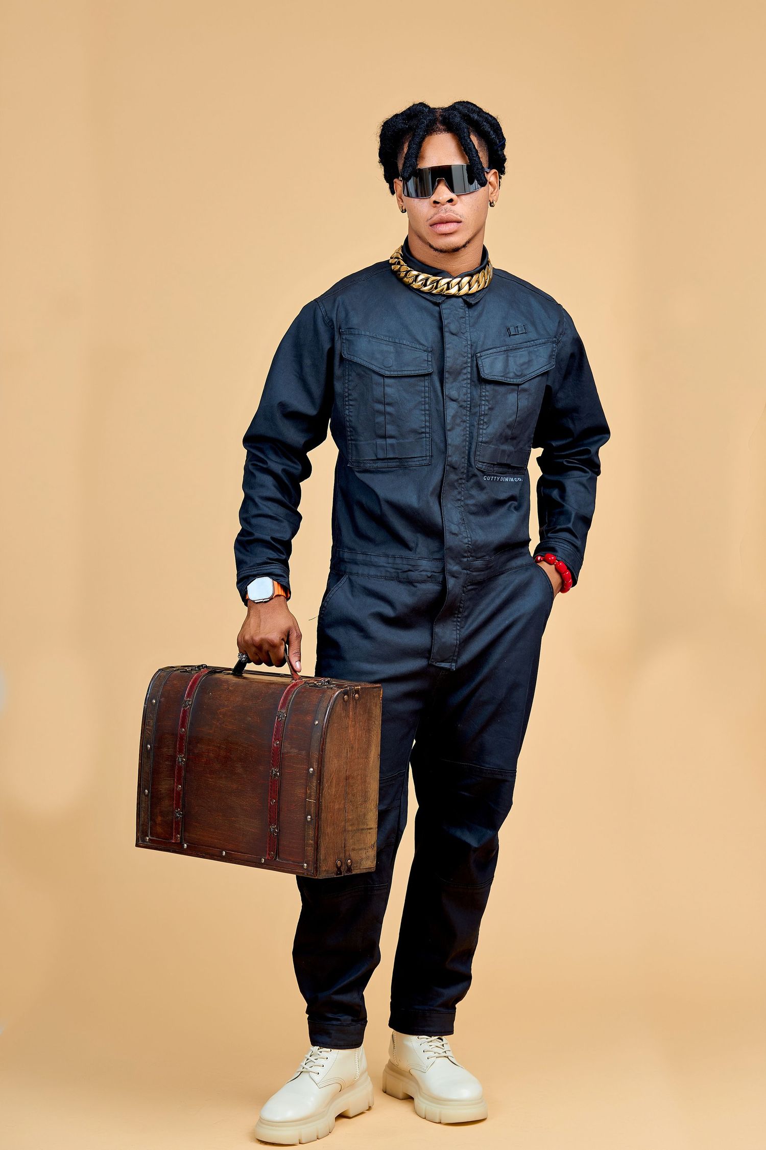 Stylish Black man in navy blue jumpsuit with gold chain holds vintage brown suitcase on beige background.