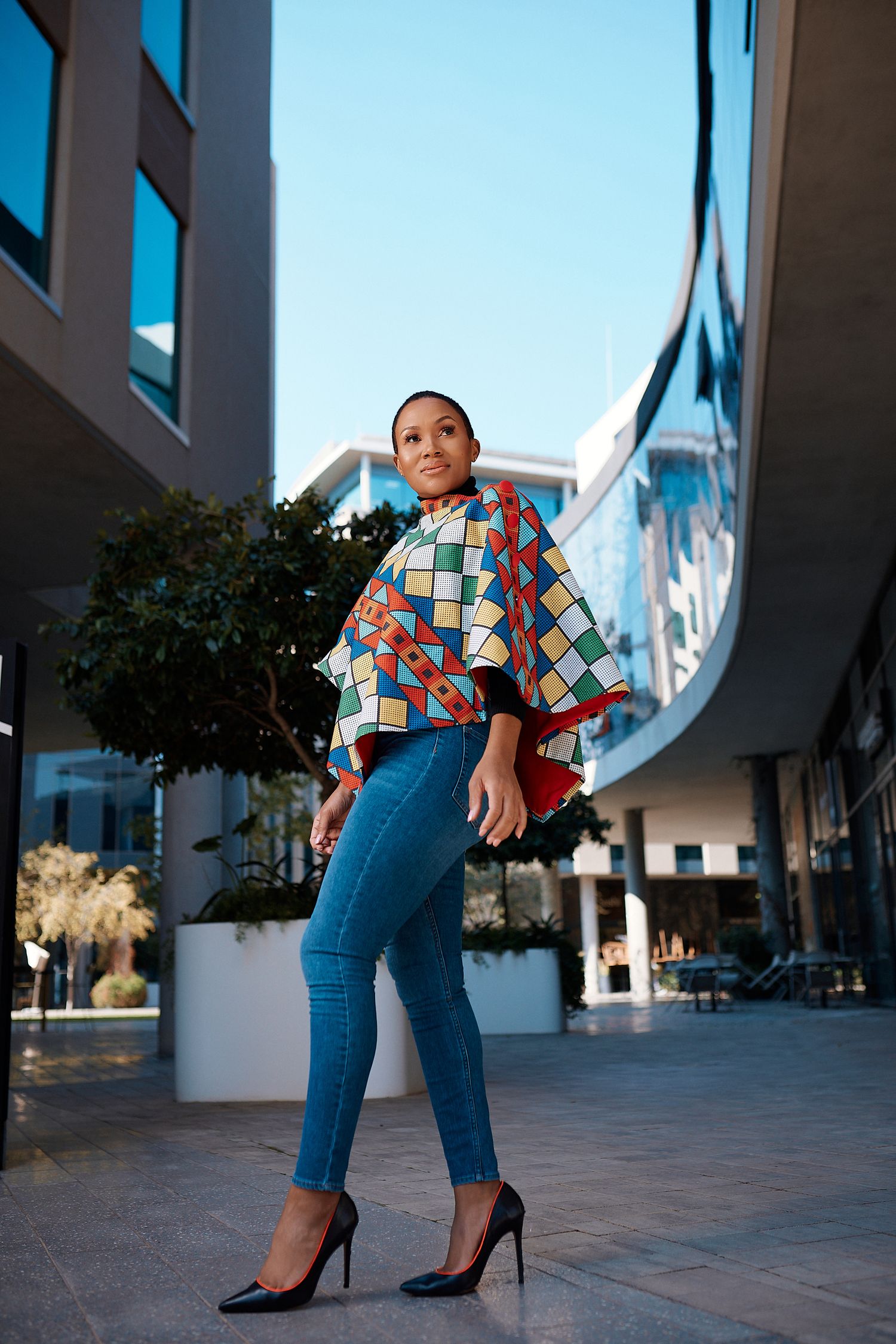Woman in colorful African print top with blue jeans and black heels posing outside building.