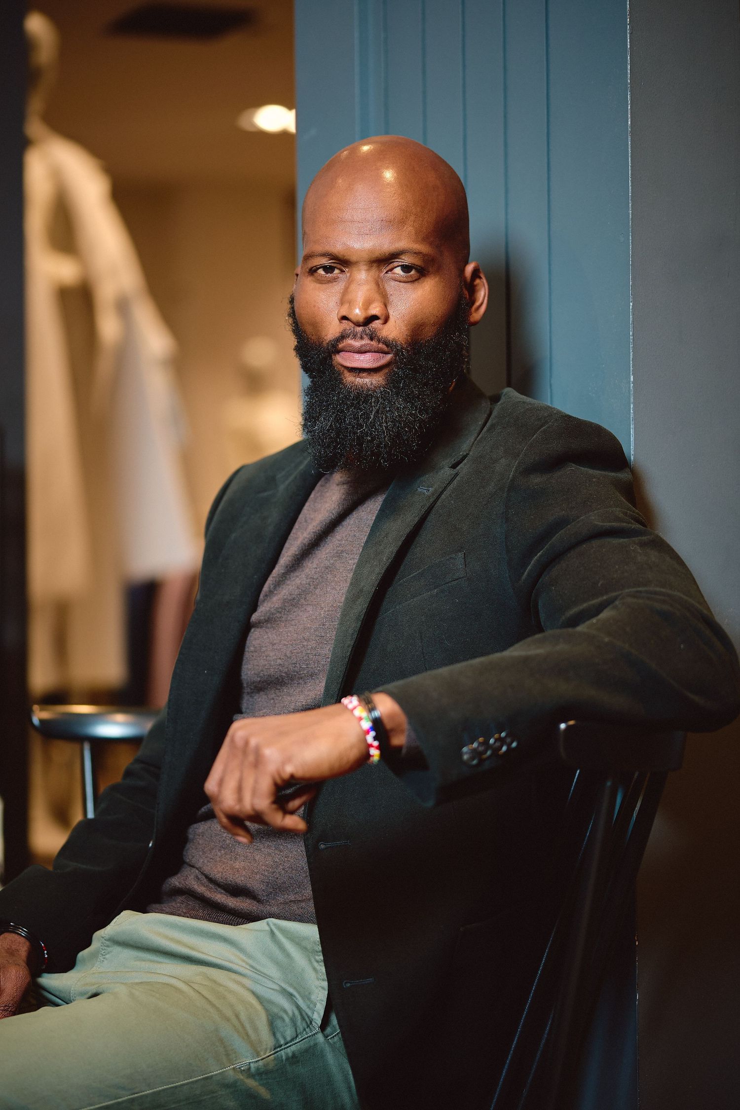 Bald Black man with full beard wearing dark blazer seated confidently in chair inside boutique store.