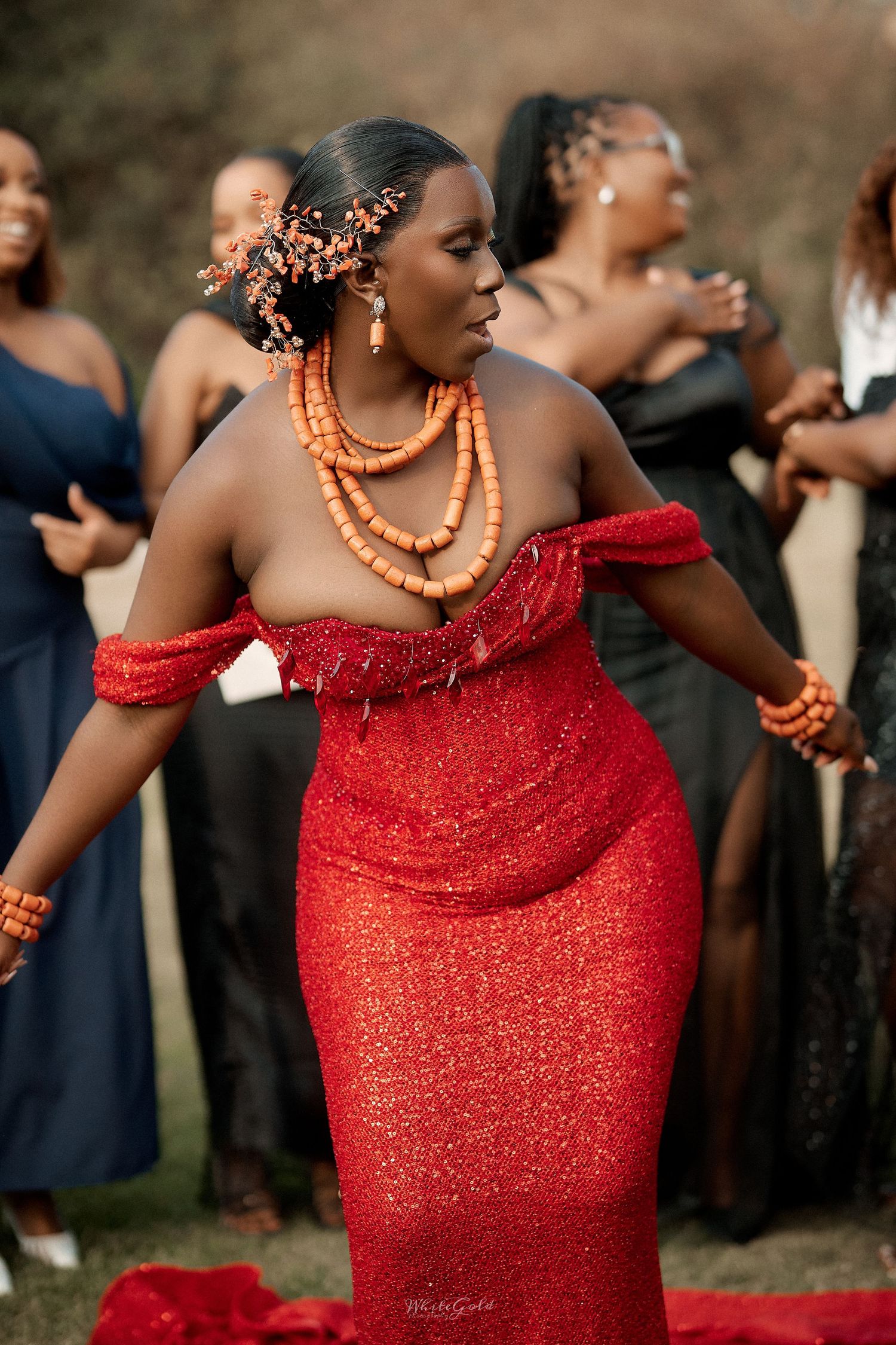 Black bride in stunning red off-shoulder glitter gown with coral beaded jewelry at outdoor wedding.