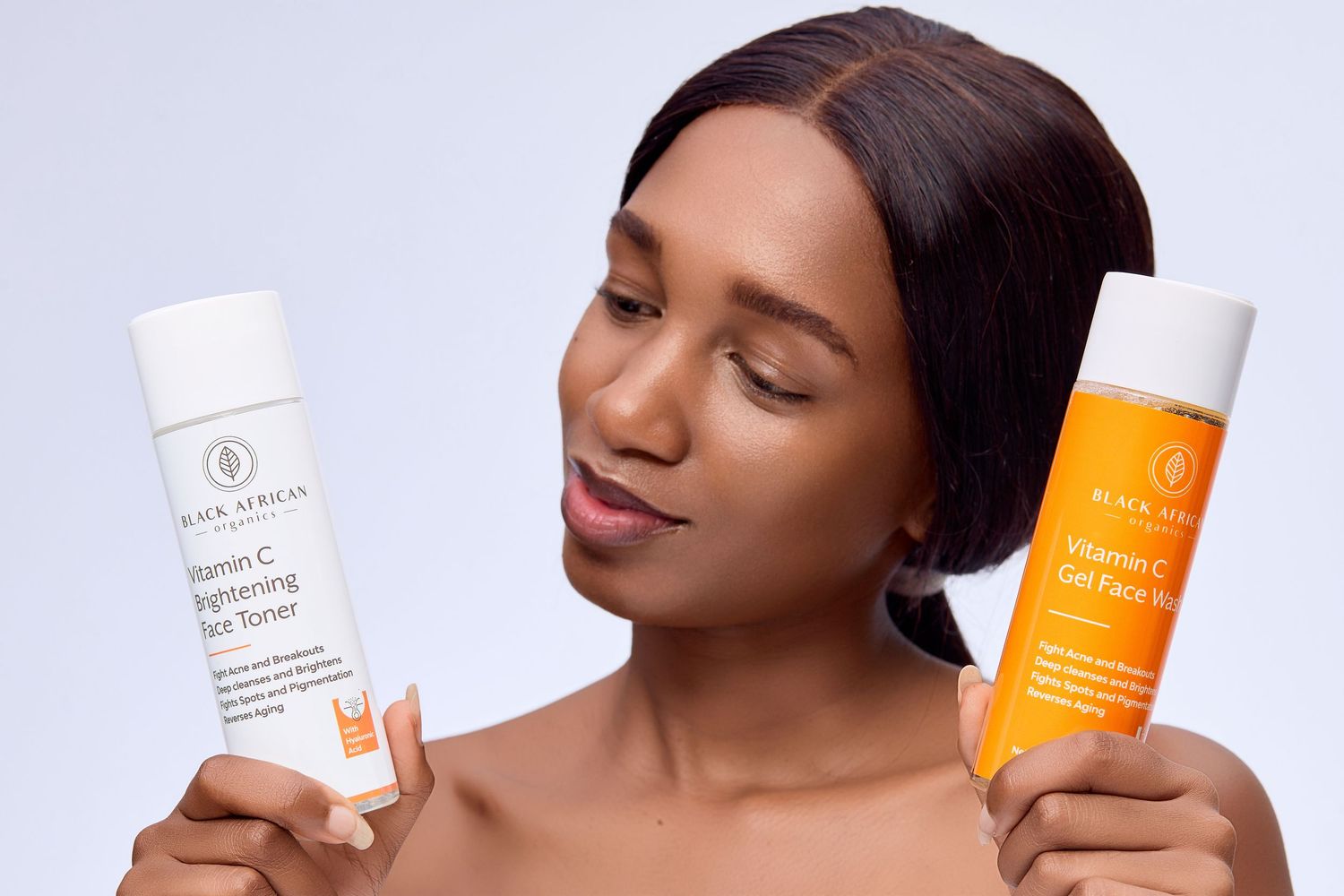 Woman holding two Vitamin C skincare products, a toner and face wash, against grey background.