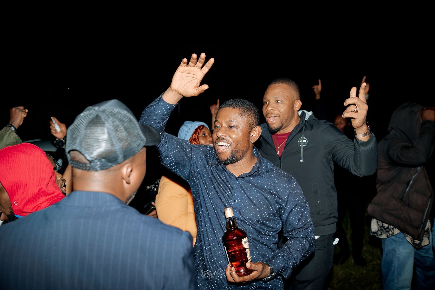 Man celebrates at night holding bottle at lively outdoor evening gathering.