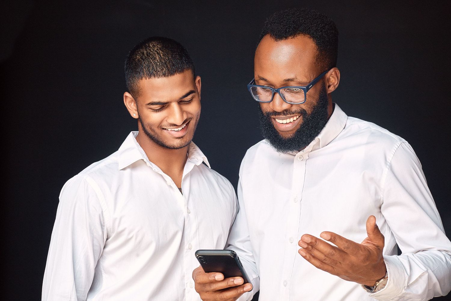 Two men in white shirts smiling while looking at a smartphone against a dark background.