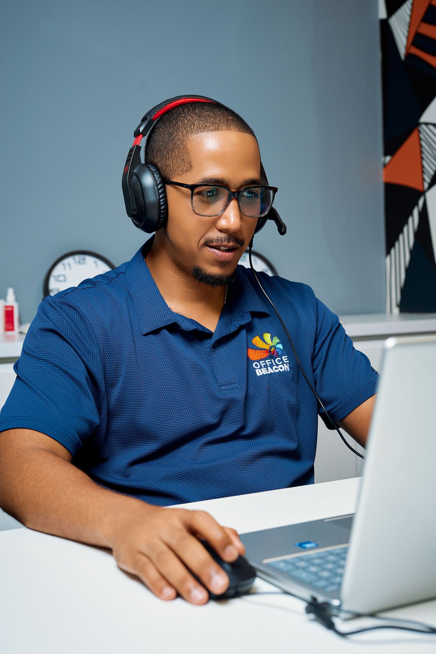 Man wearing headset and glasses works on laptop at Office Beacon customer service desk.