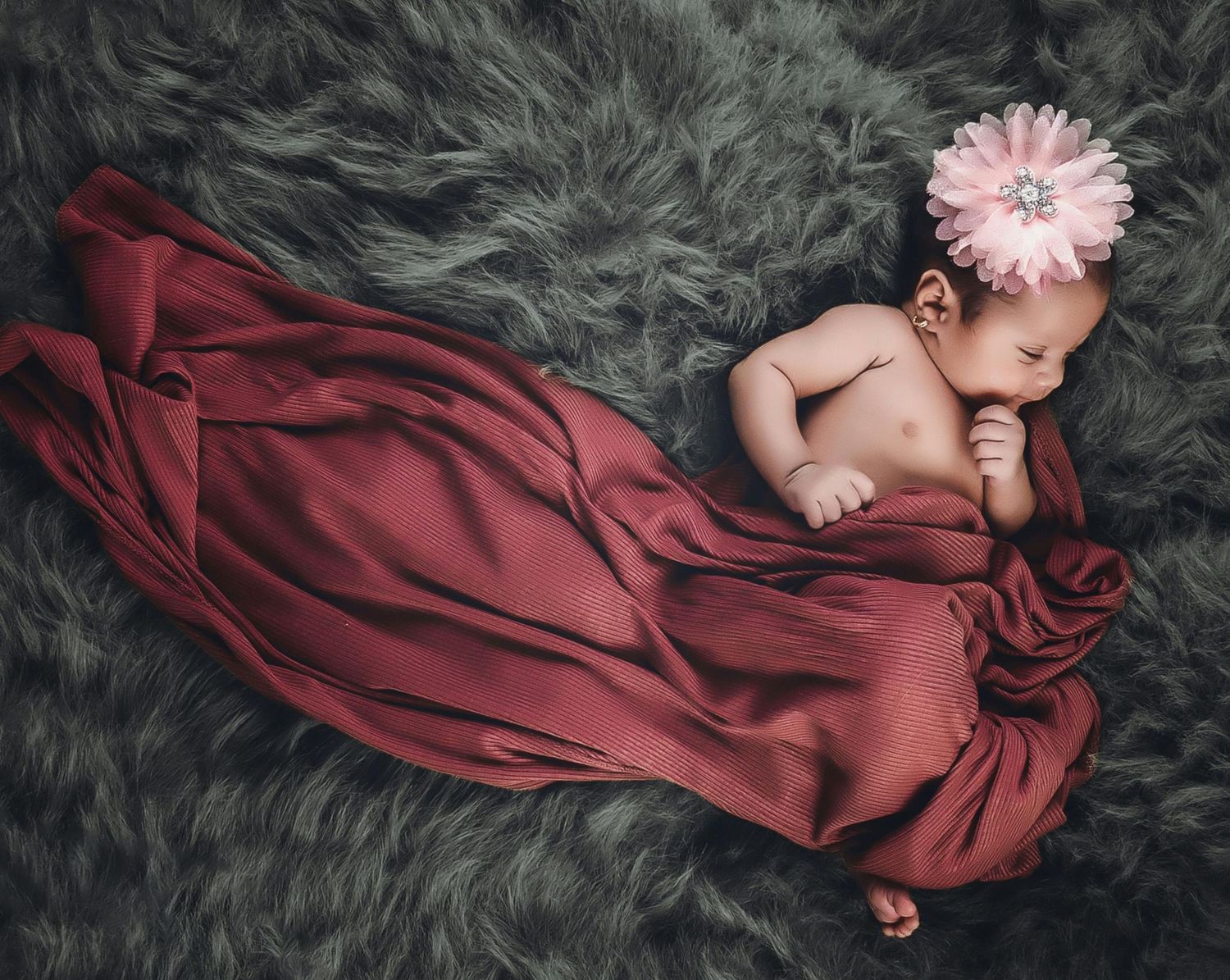Sleeping newborn baby wrapped in burgundy fabric with pink floral headband on gray fluffy blanket.