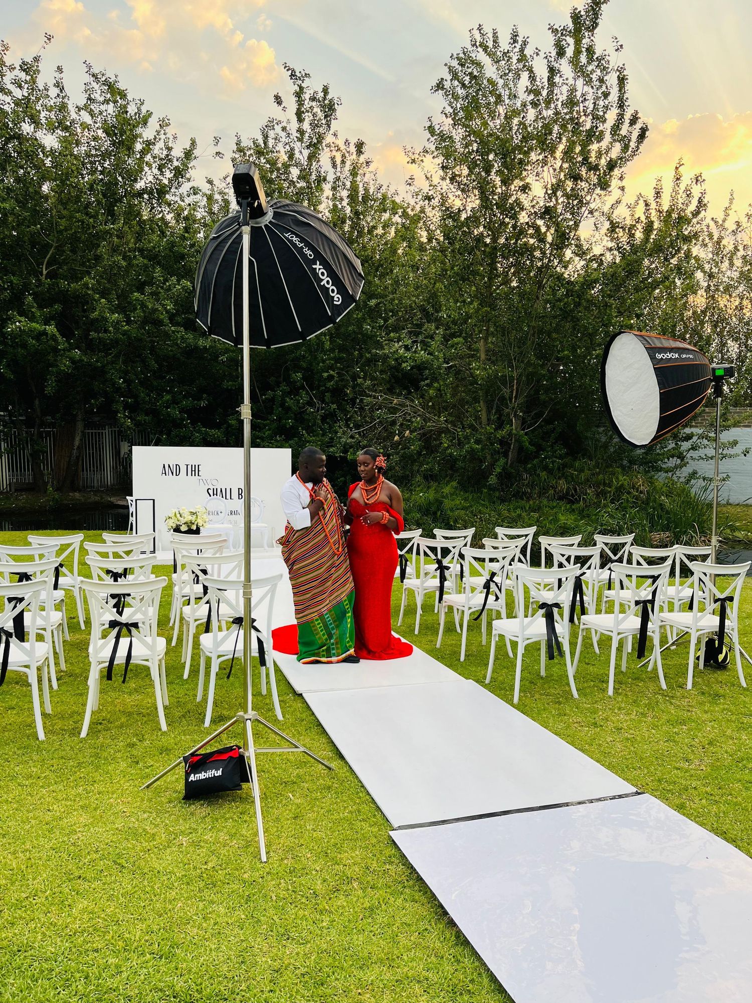 Outdoor wedding ceremony setup at sunset with white chairs, aisle, and two guests in red attire posing.