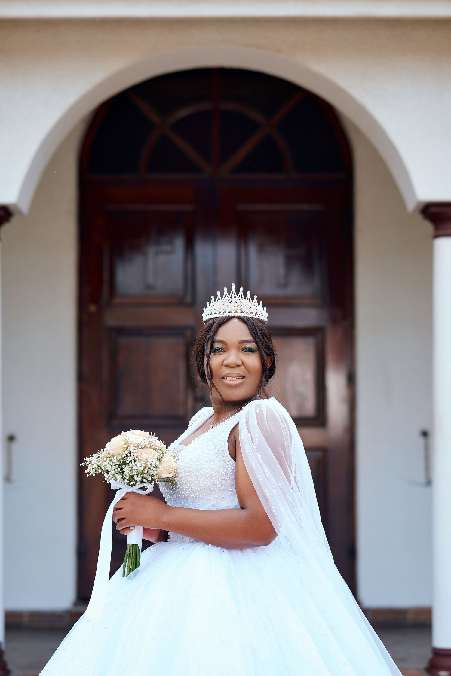 Elegant Black bride in white ball gown with pearl details, crown tiara, and bouquet at church entrance.