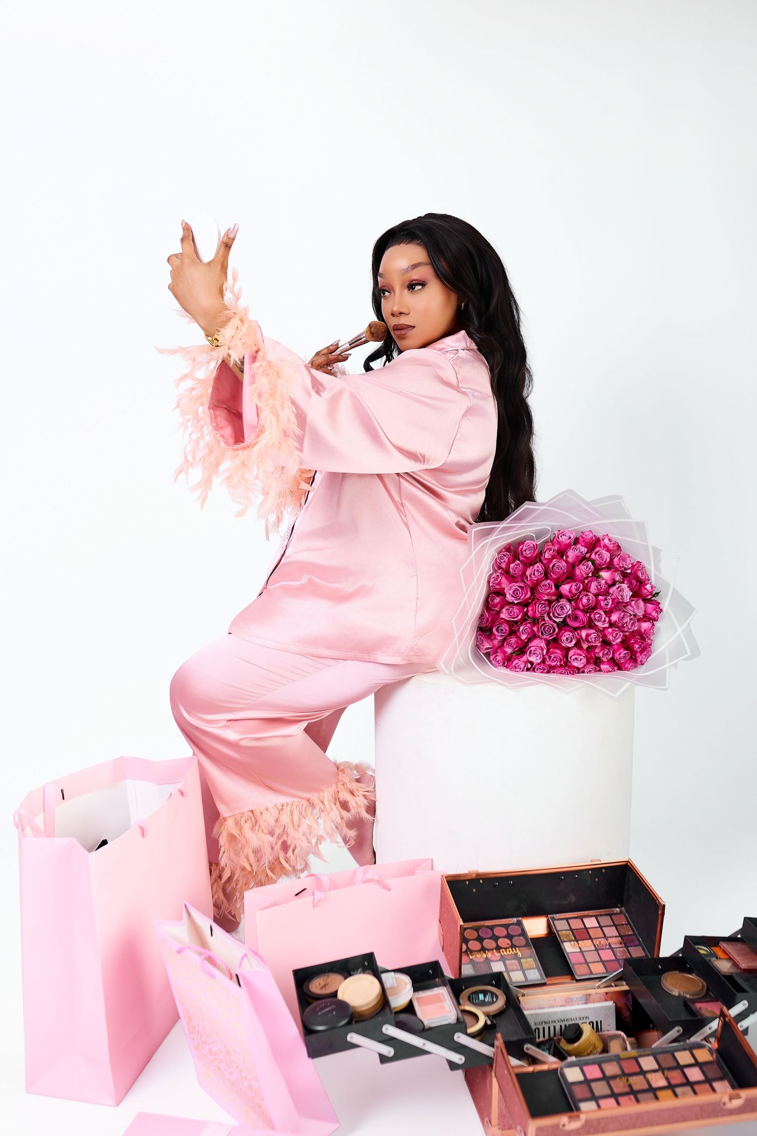 Stylish Black woman in pink feather-trimmed satin suit posing with roses, makeup palettes and pink shopping bags.