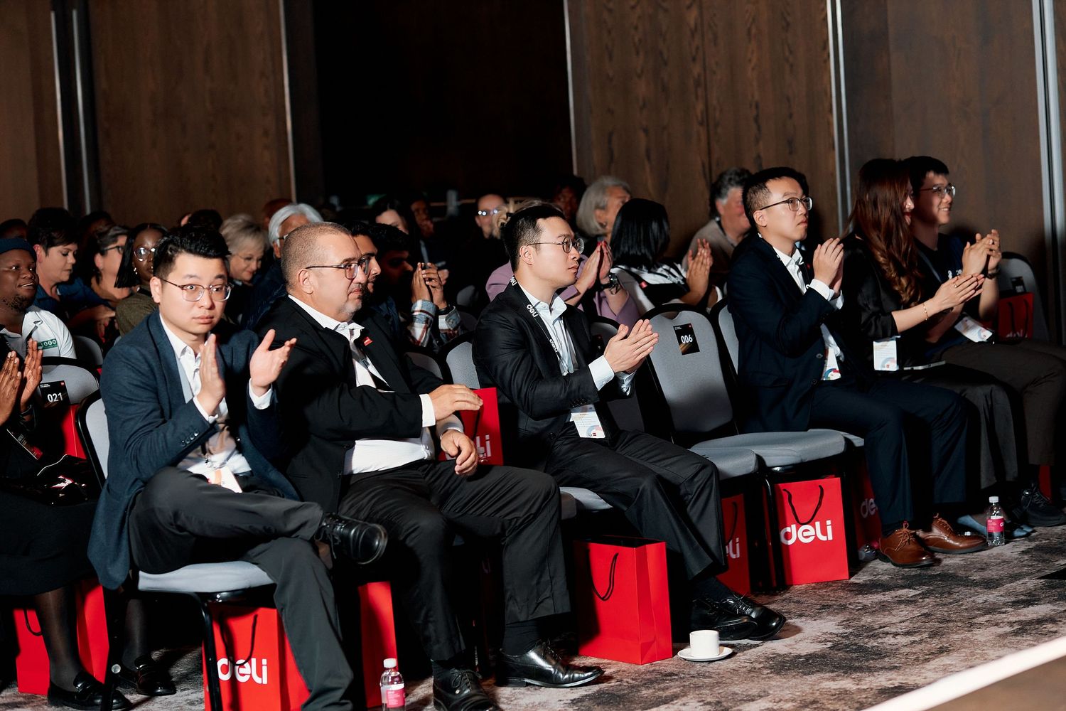 Business professionals in formal attire applaud at a corporate event, seated in rows at a conference venue.