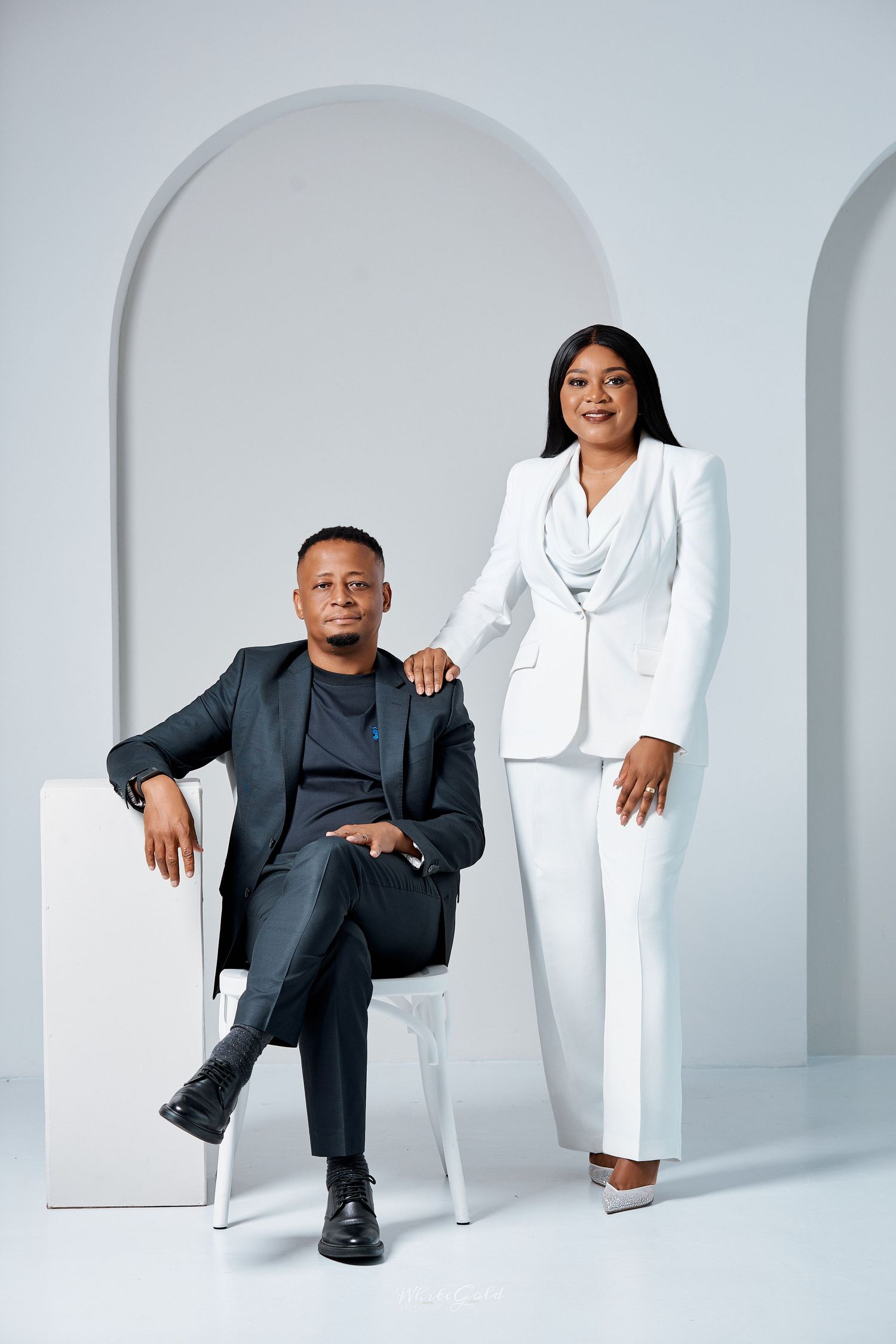 A Black man in a dark suit sits beside a Black woman in a white pantsuit in an elegant studio setting.