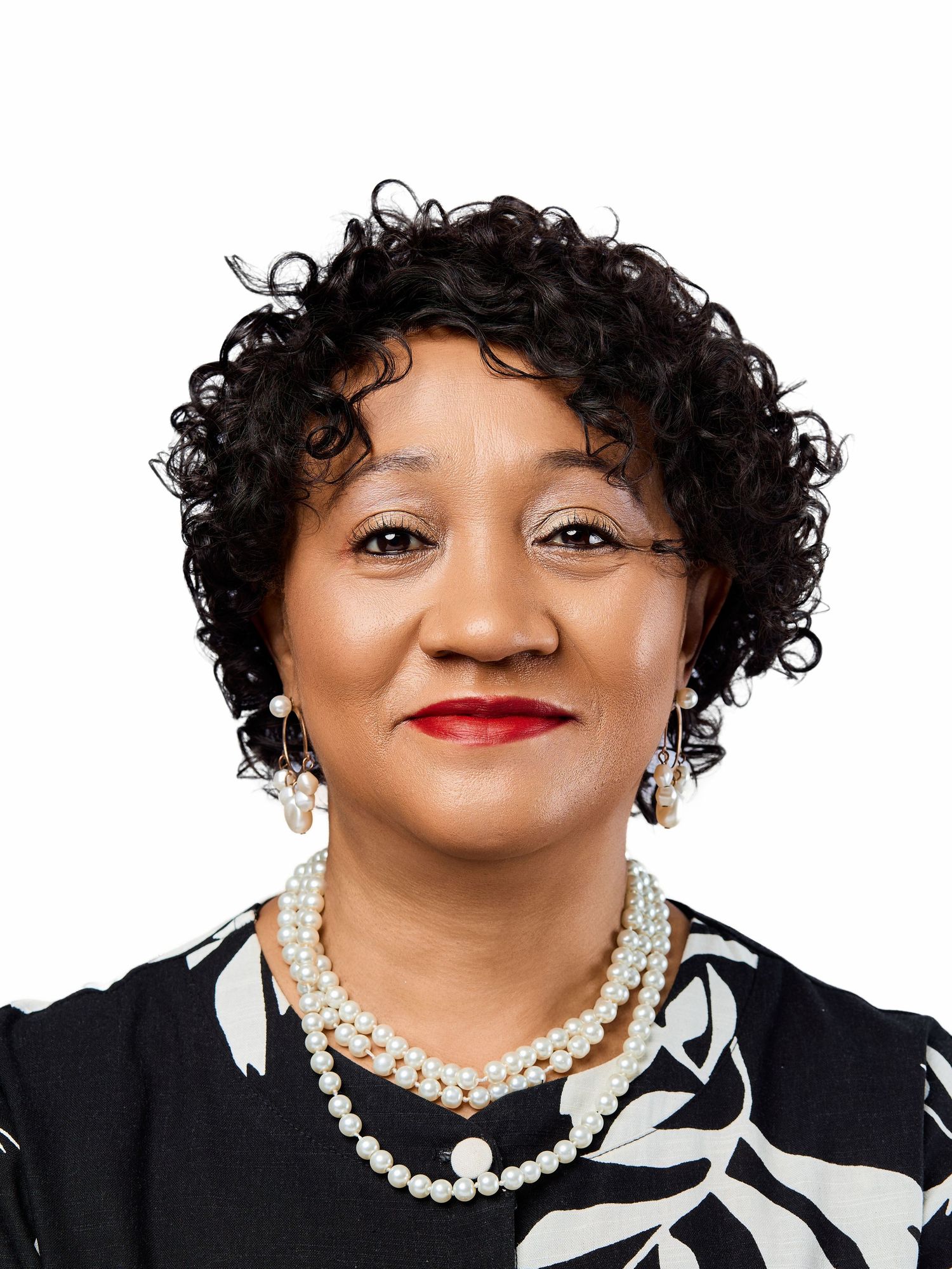 Professional headshot of a smiling woman with curly hair wearing pearls and a floral black top.