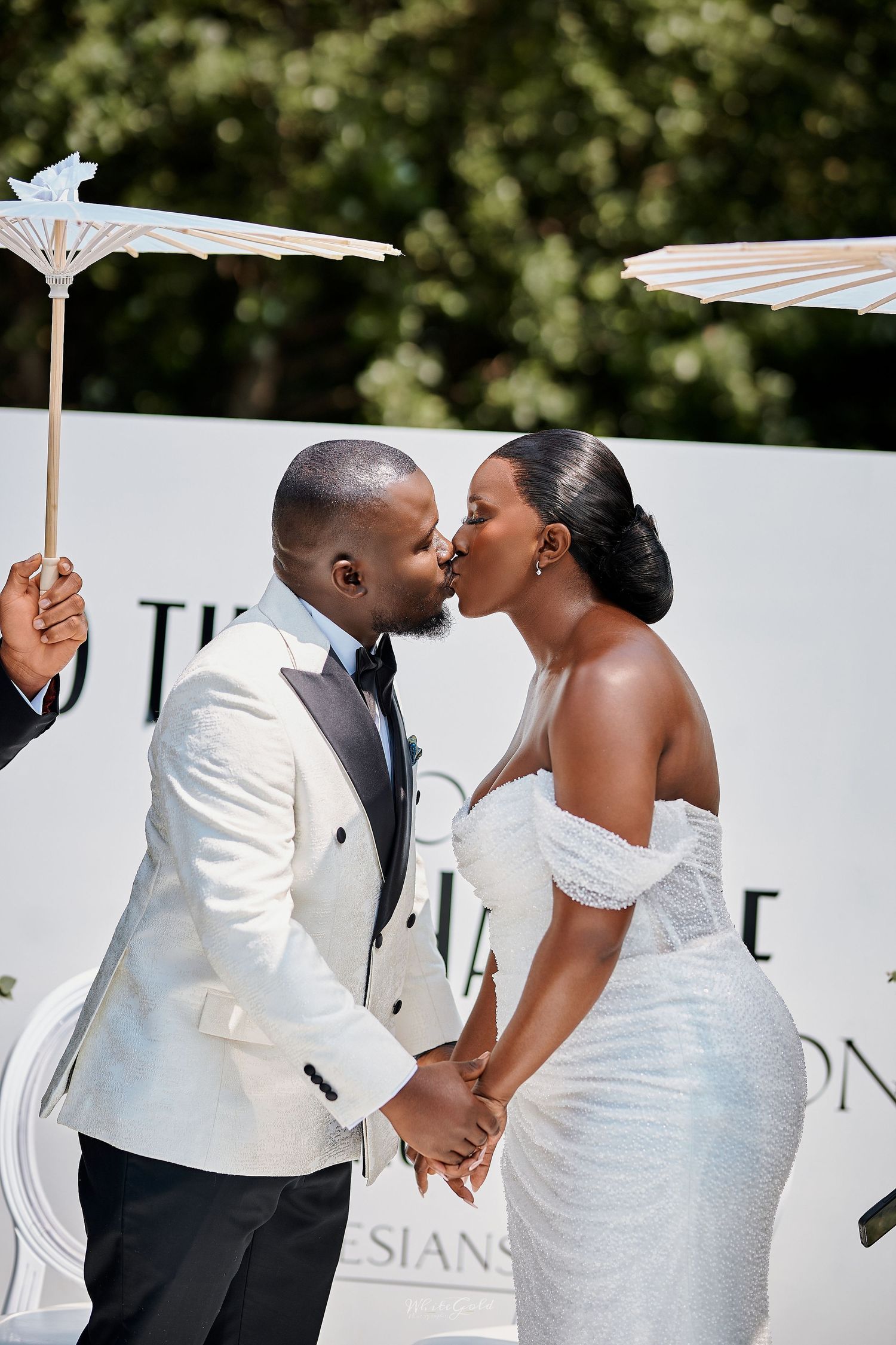 Couple sharing a kiss at outdoor wedding ceremony under white parasol in sunlight.