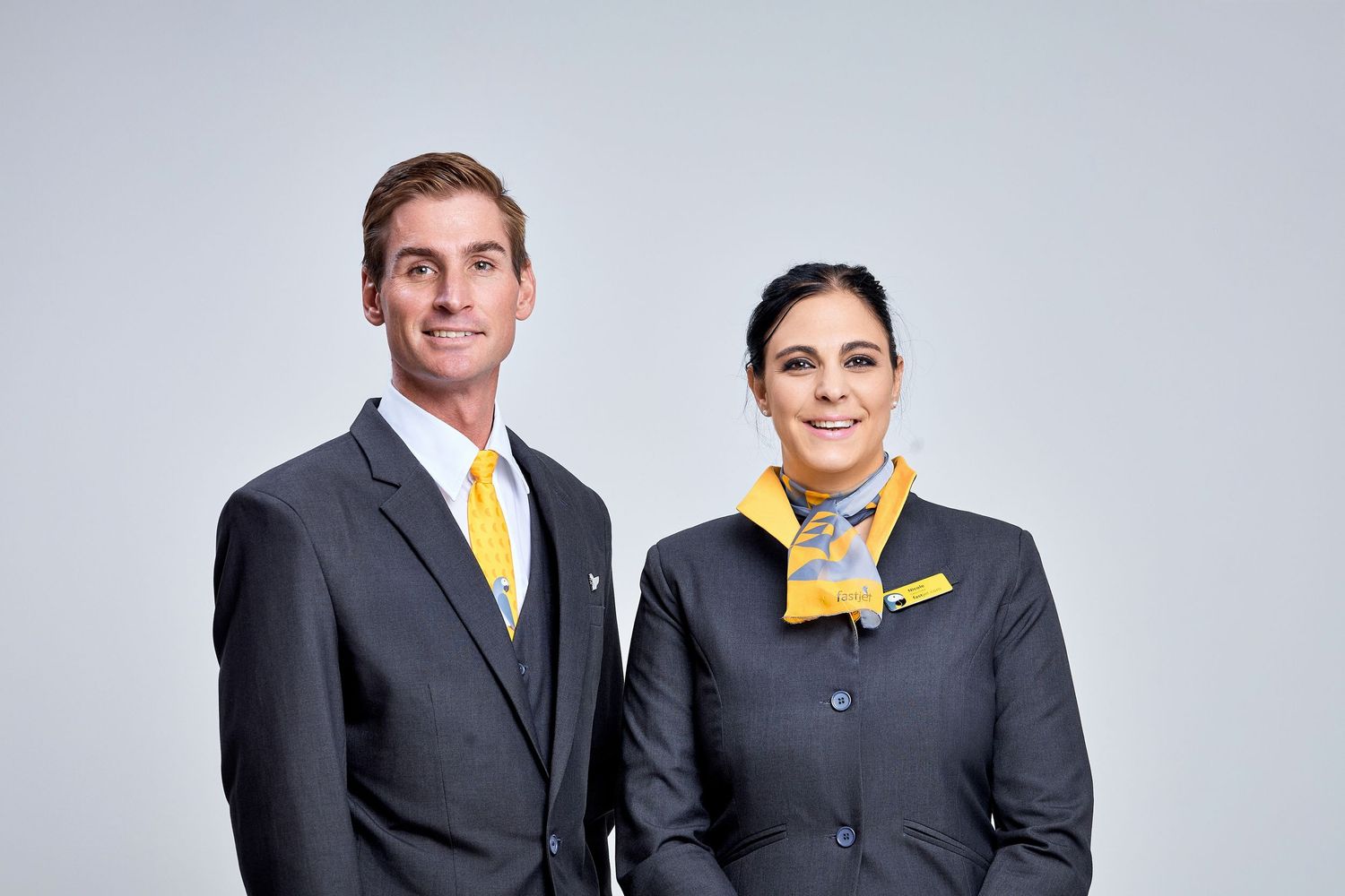 Male and female airline staff in matching dark gray uniforms with yellow ties posing professionally.