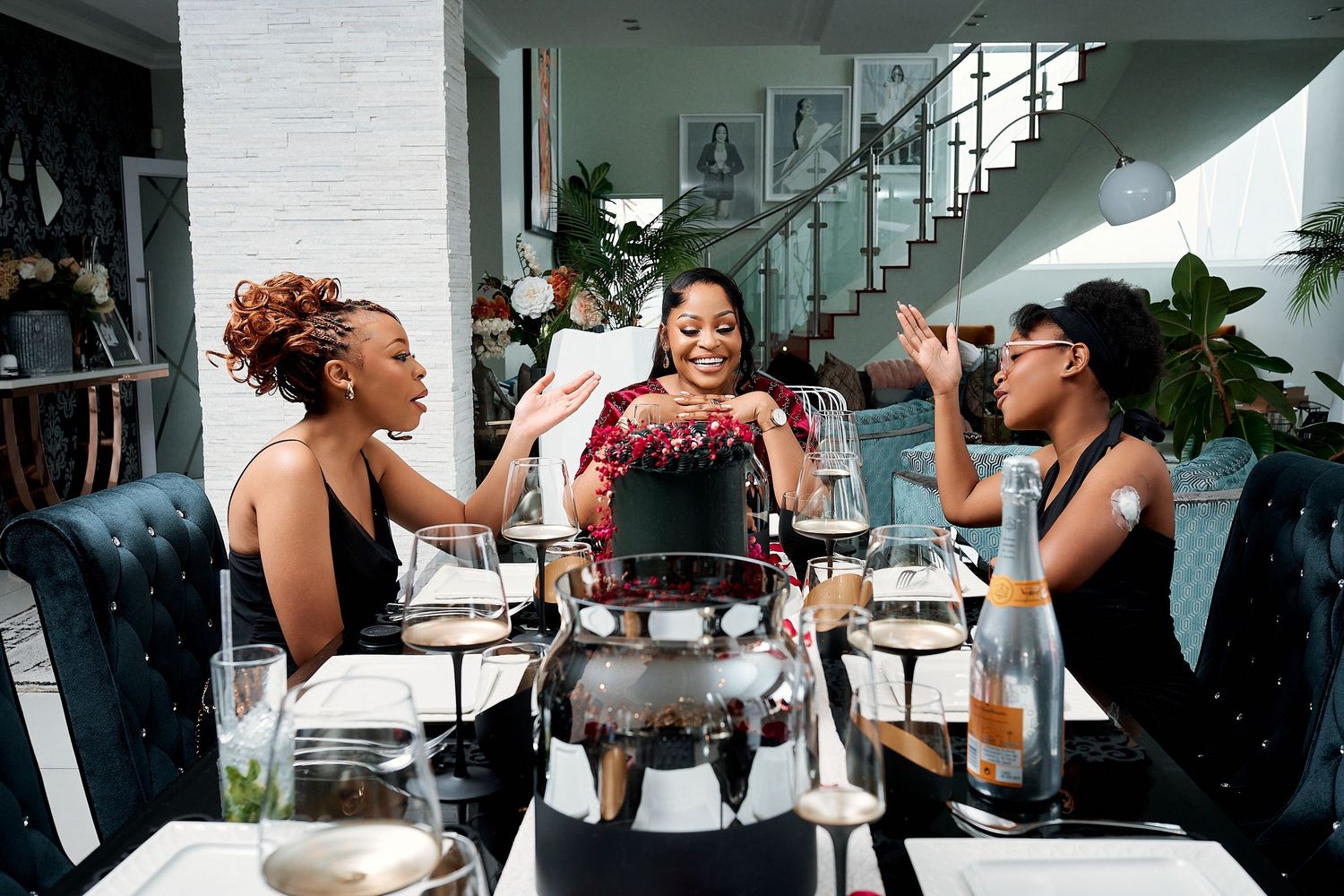 Three women laugh and celebrate together at a stylishly set dining table indoors.
