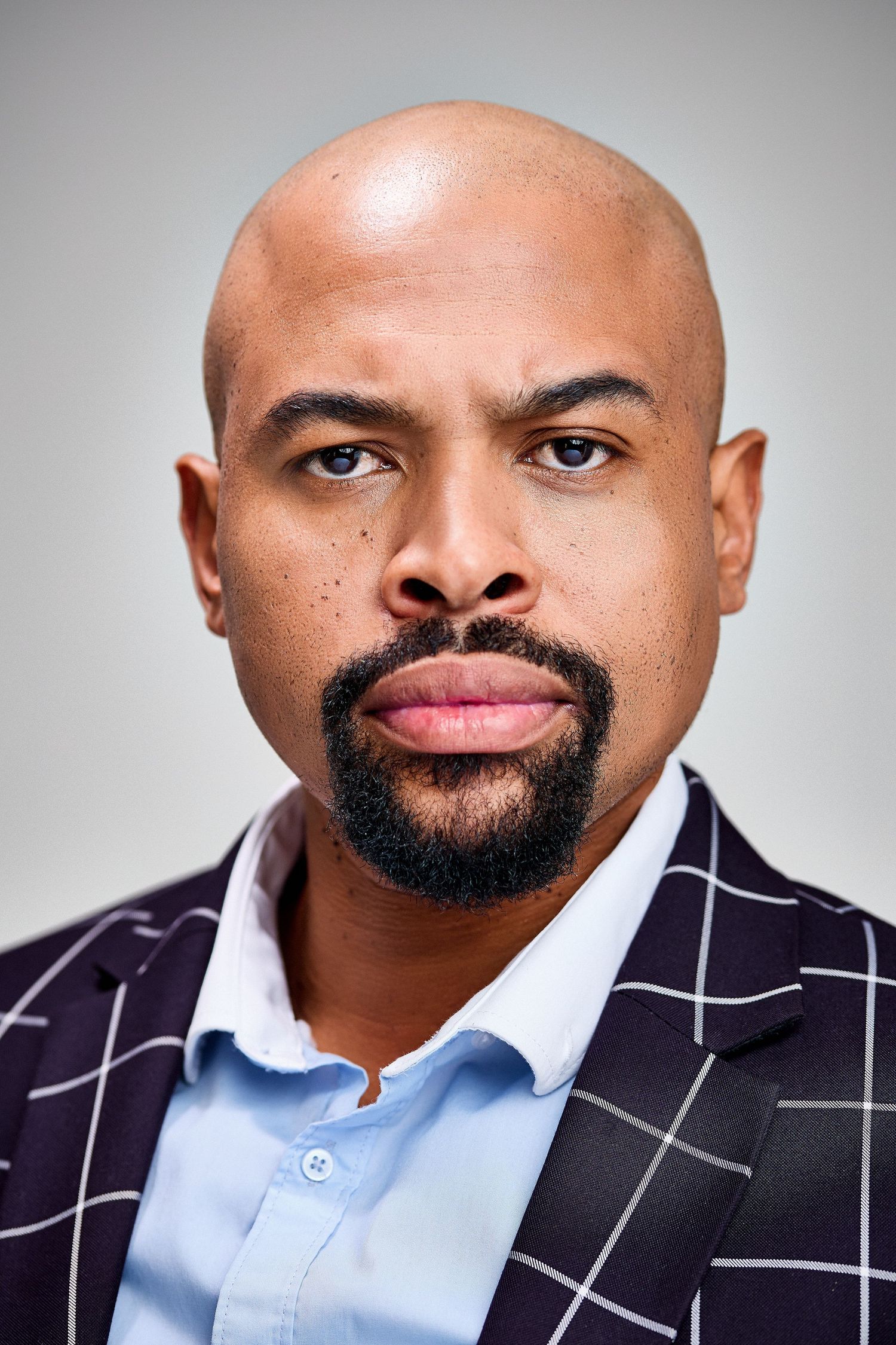 Bald Black man with beard wearing a plaid blazer and light blue shirt poses for a professional headshot.
