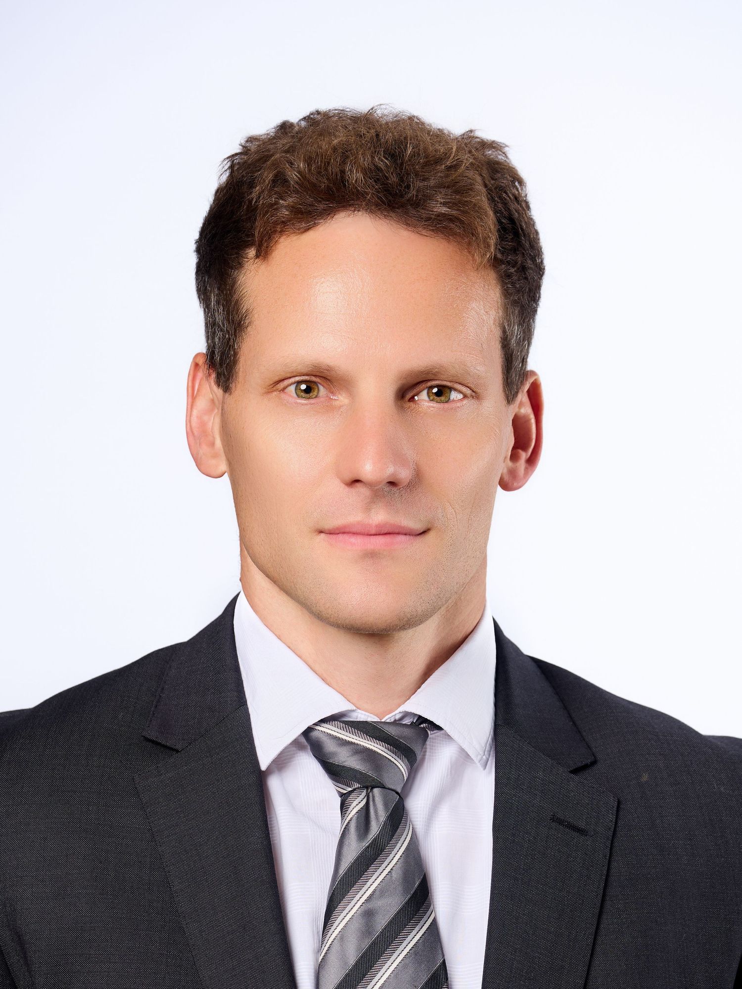 Professional headshot of a man with brown curly hair in a dark suit and striped tie against a white background.