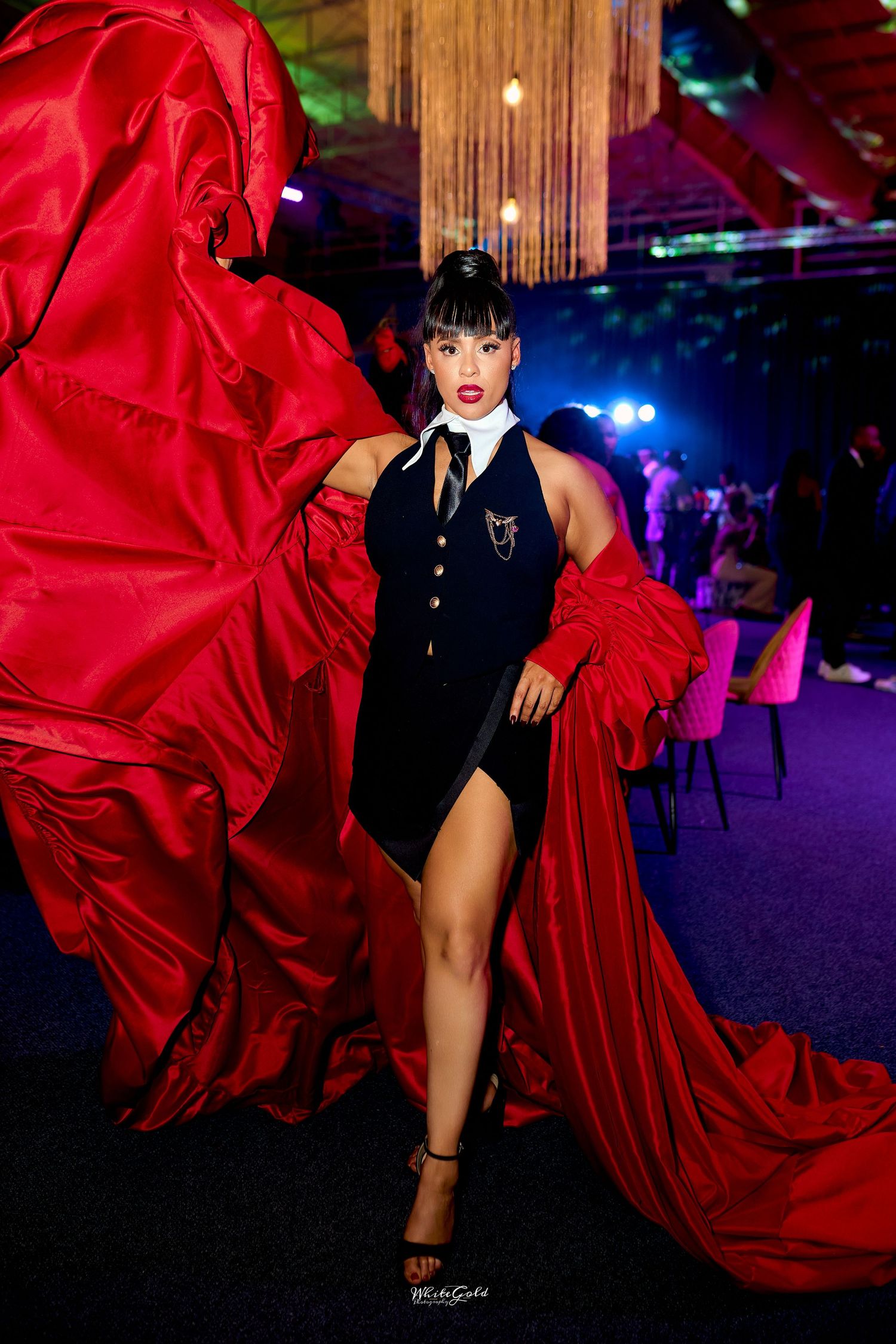 Woman in black halter bodysuit with dramatic red flowing cape at glamorous evening event.
