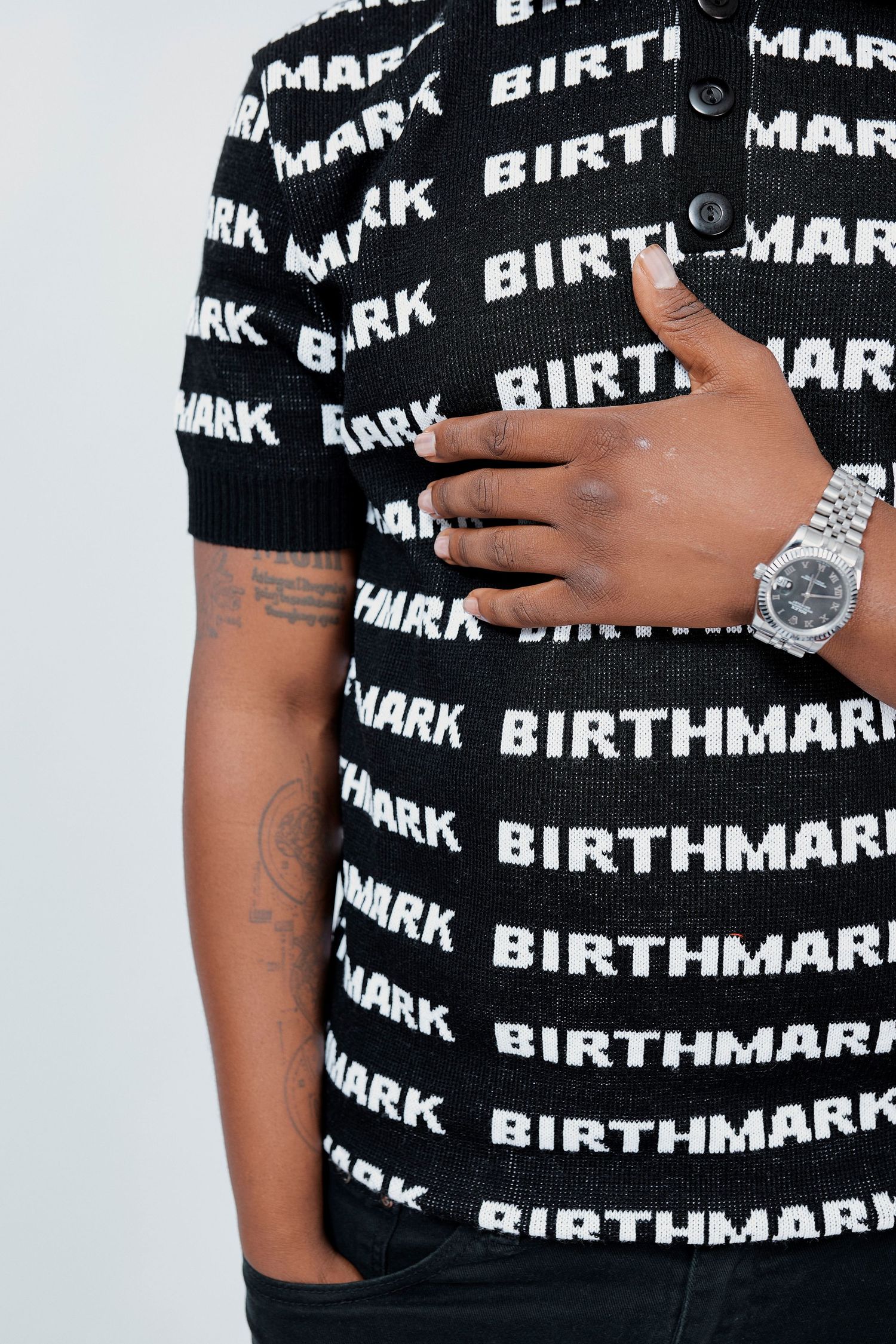 Close-up of man in black Birthmark logo knit polo shirt wearing a silver luxury watch.