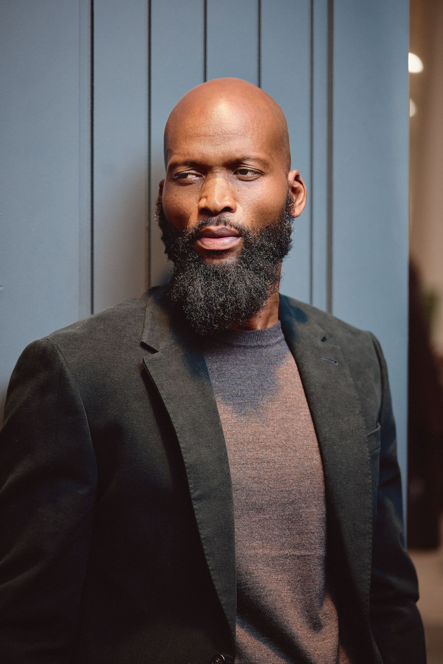 Bald Black man with full beard wearing dark blazer poses against blue paneled wall background.