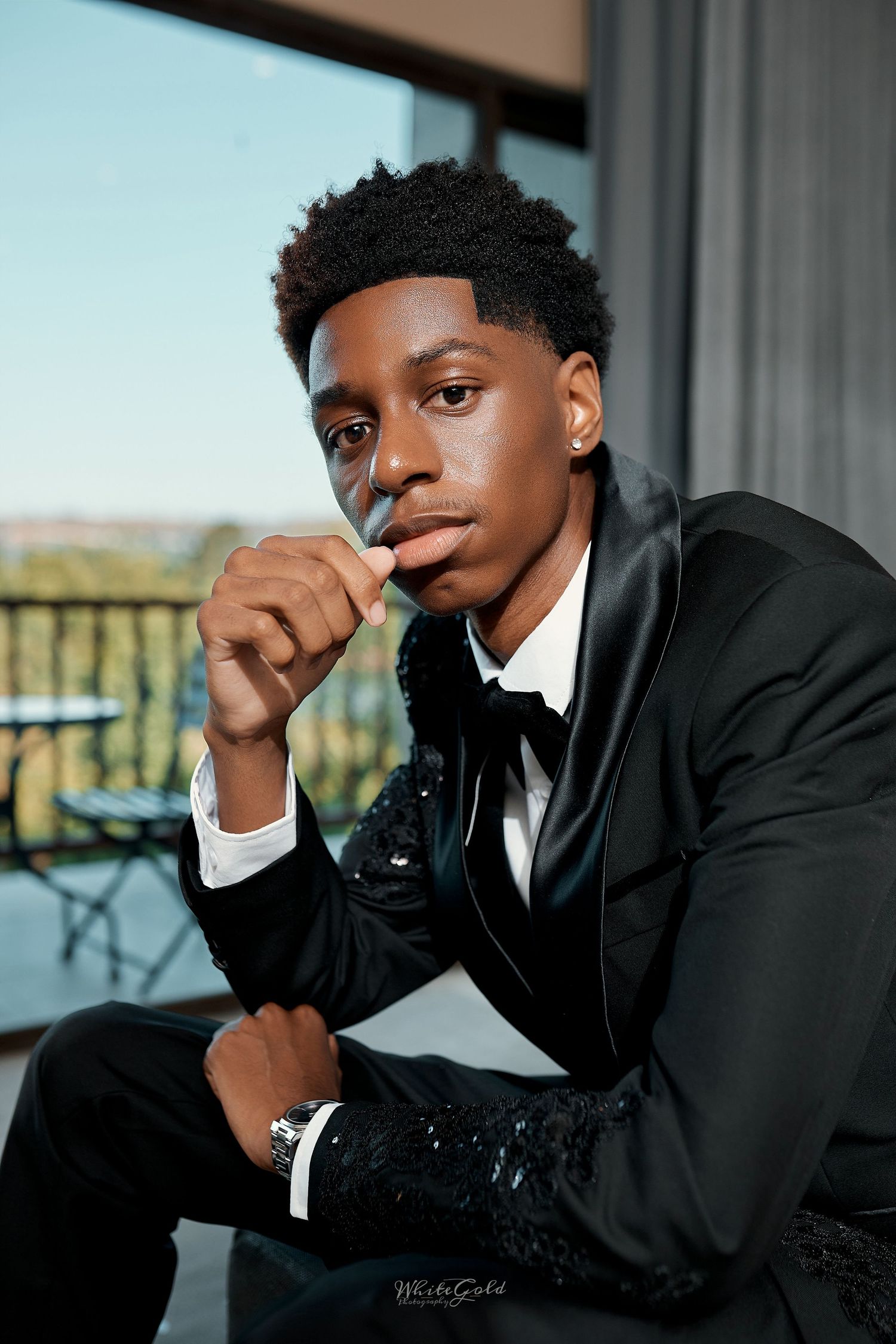 Young Black man in black sequin tuxedo sits pensively by window in professional portrait photo.