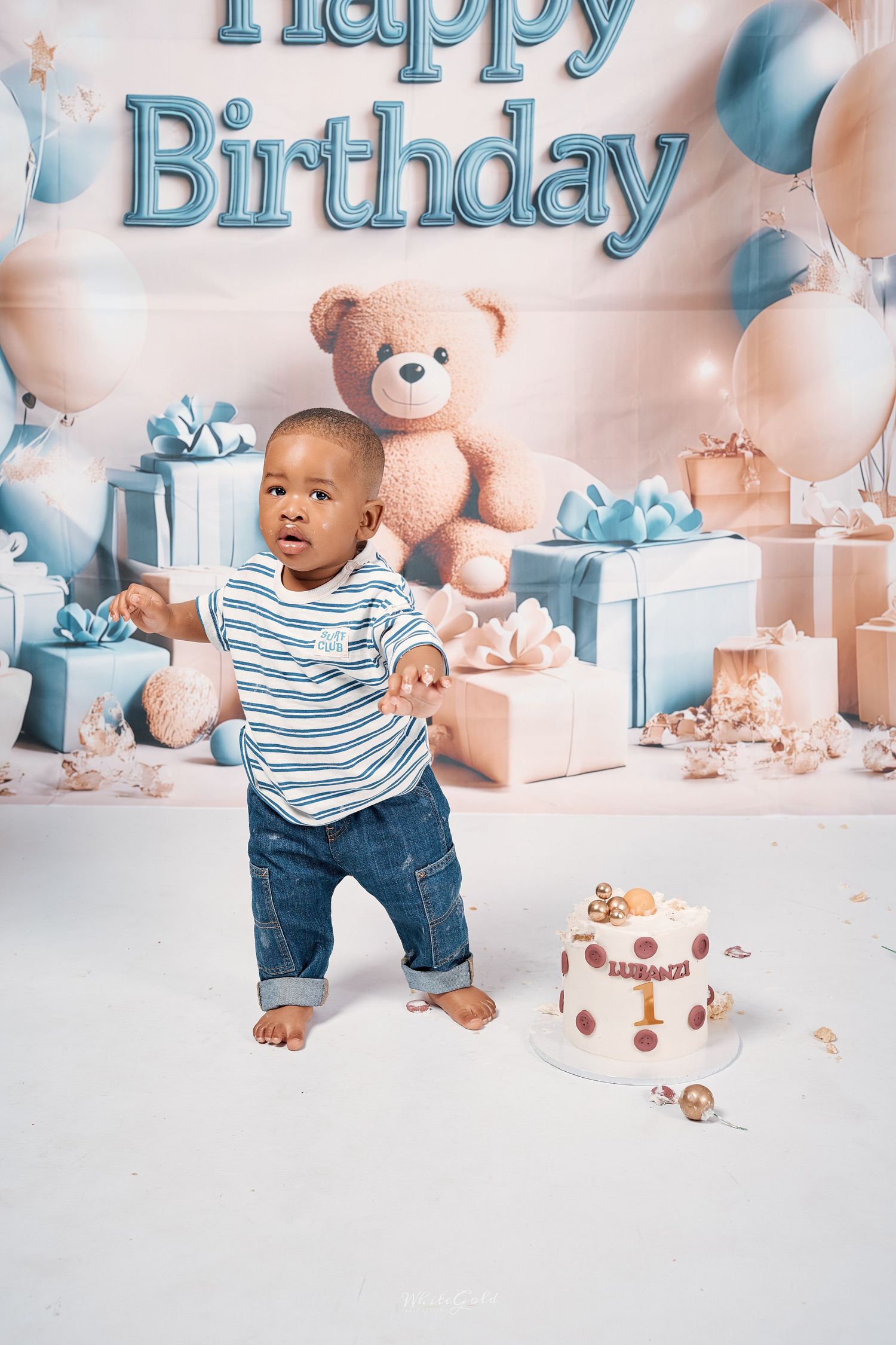 A Black toddler boy stands barefoot in jeans and striped shirt beside a birthday cake at his first birthday party.