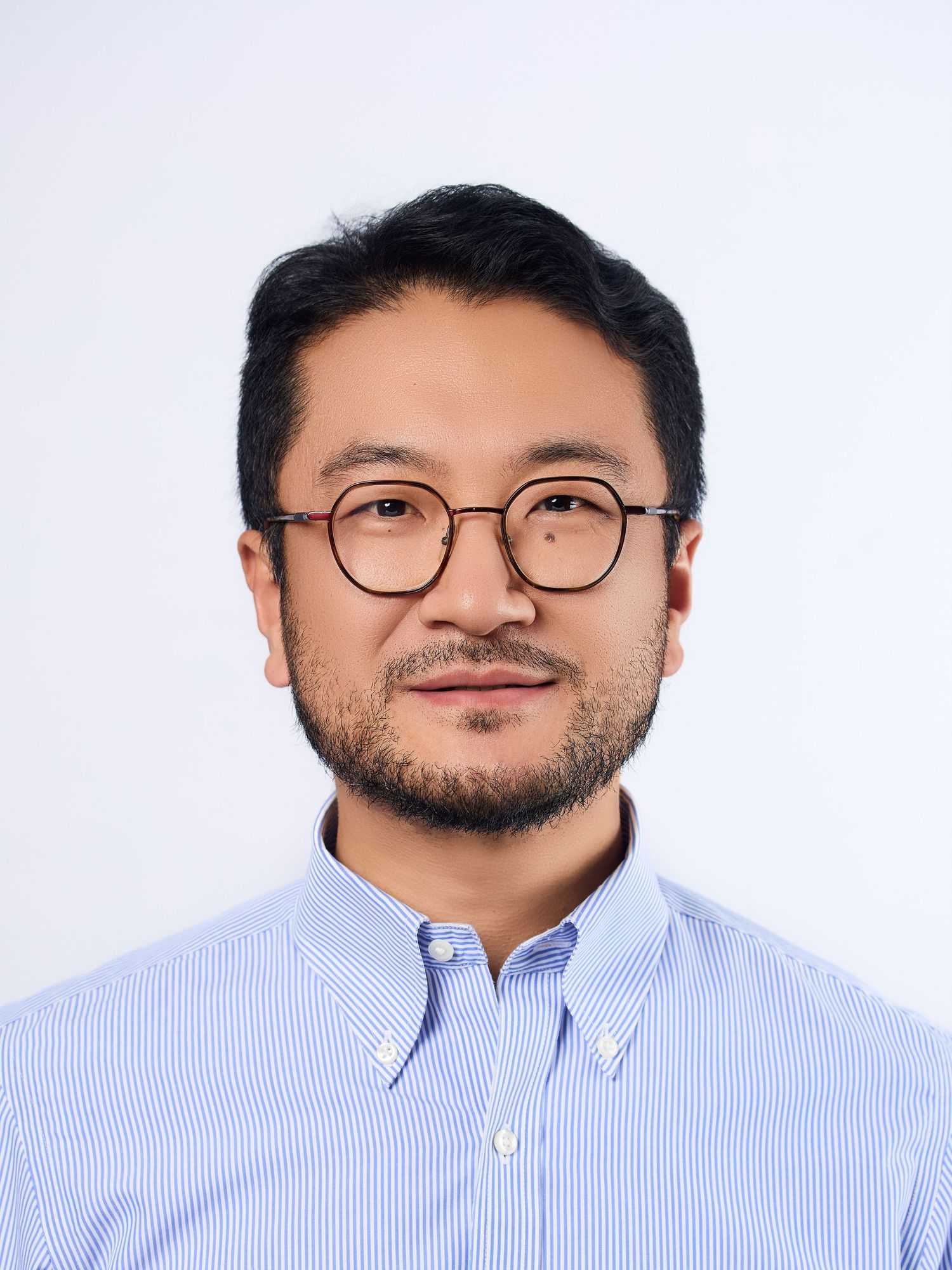 Asian man with round glasses and short dark hair wearing a light blue striped button-down shirt against white background.