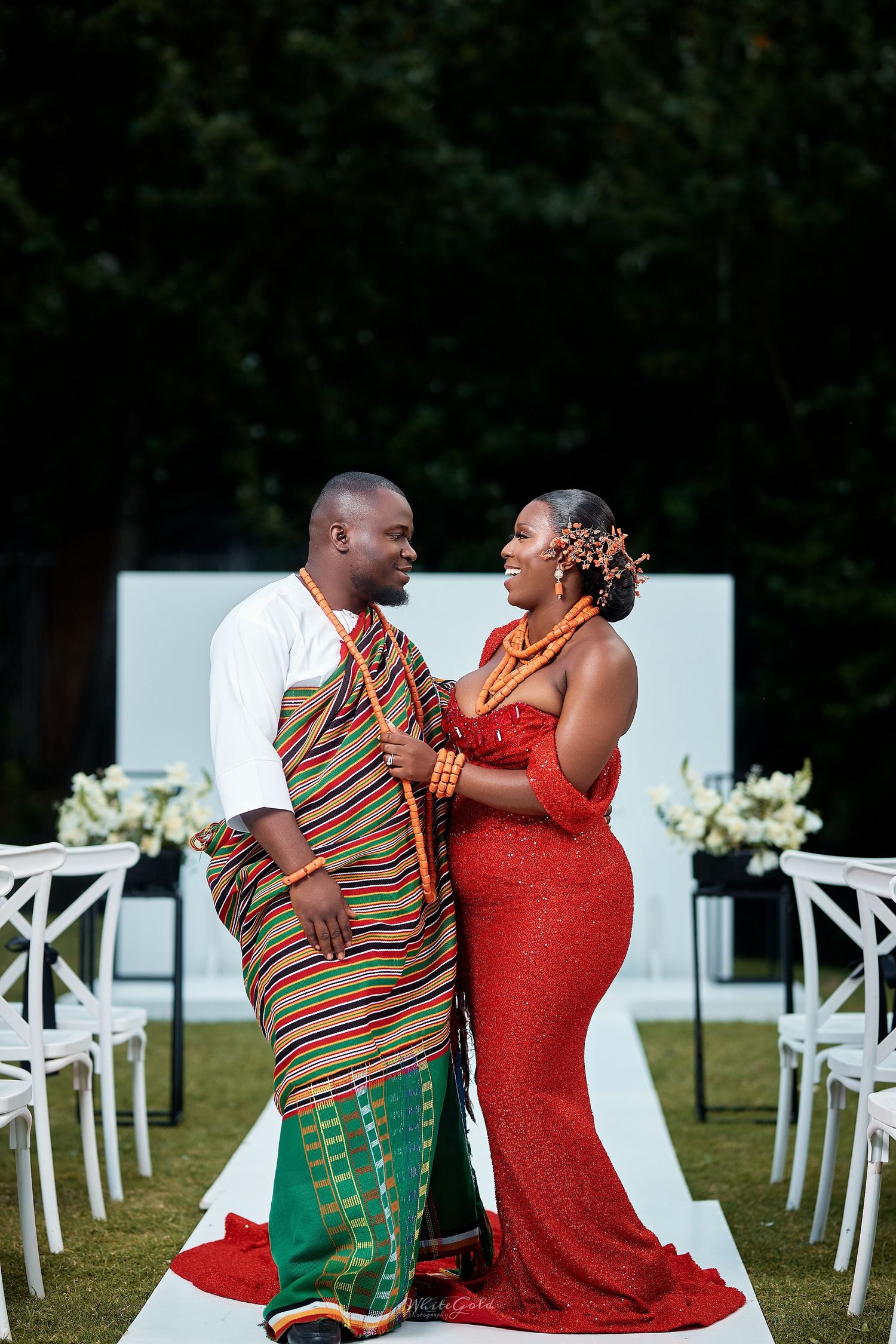 Couple in traditional African Kente and red ceremonial attire sharing romantic gaze at outdoor wedding.