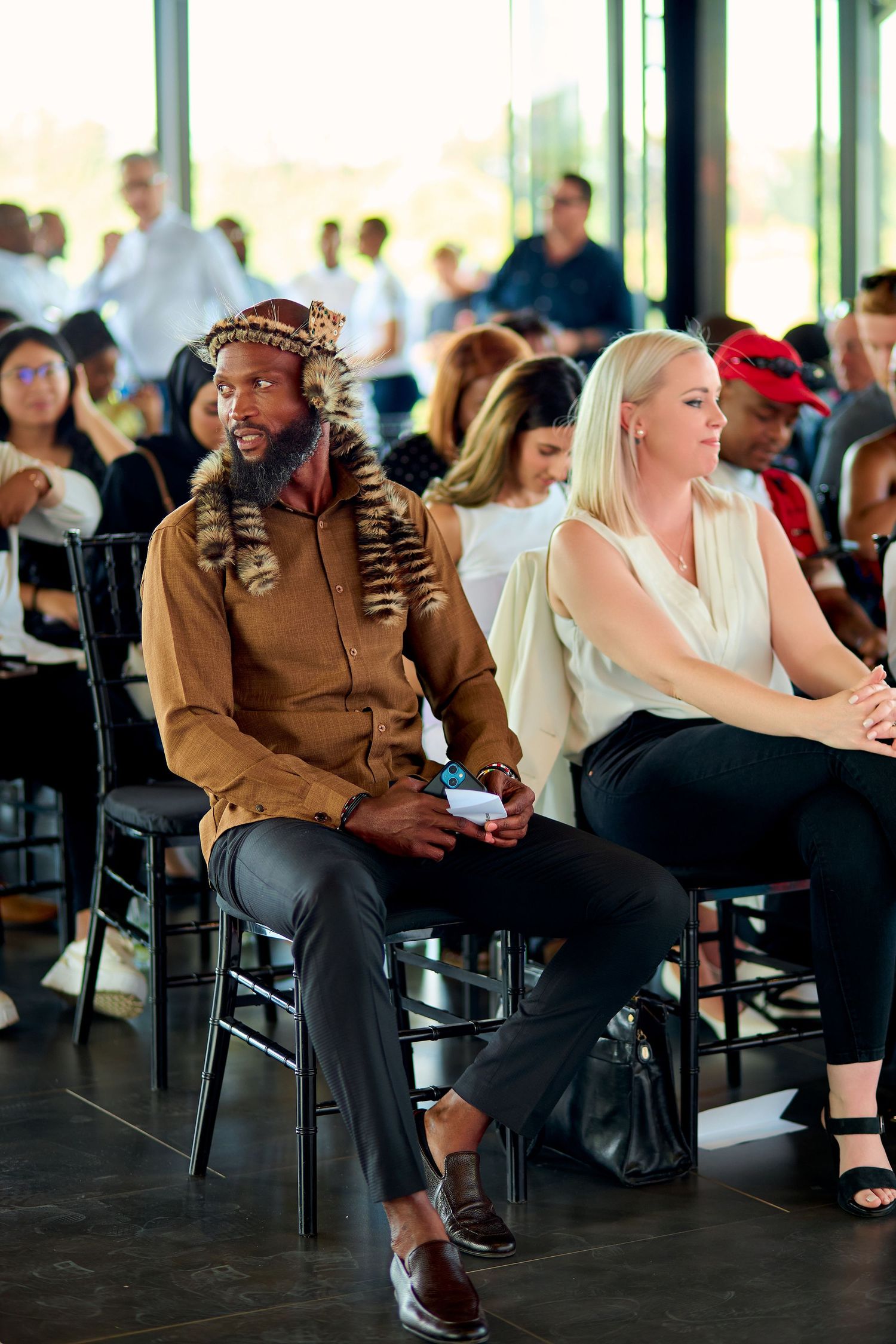 Man in traditional African leopard skin attire seated beside blonde woman at formal event.