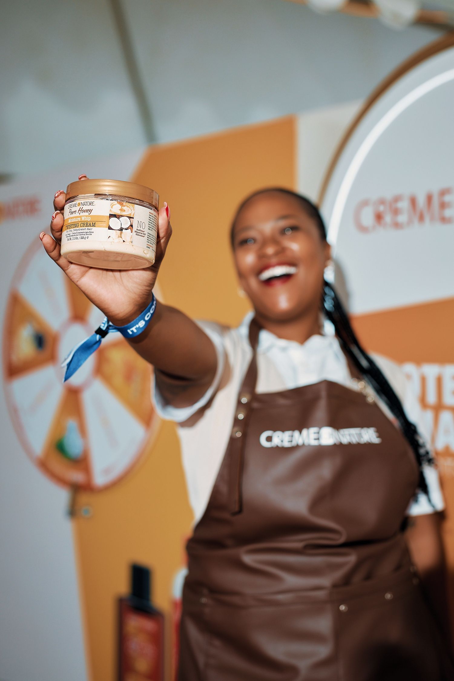 Smiling Creme of Nature brand ambassador in apron holding Pure Honey hair product at event.