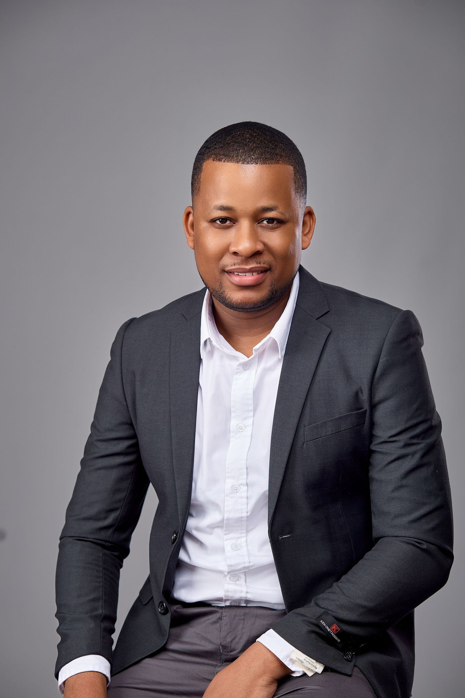 Confident Black man in dark blazer and white shirt smiles while seated against a gray studio background.