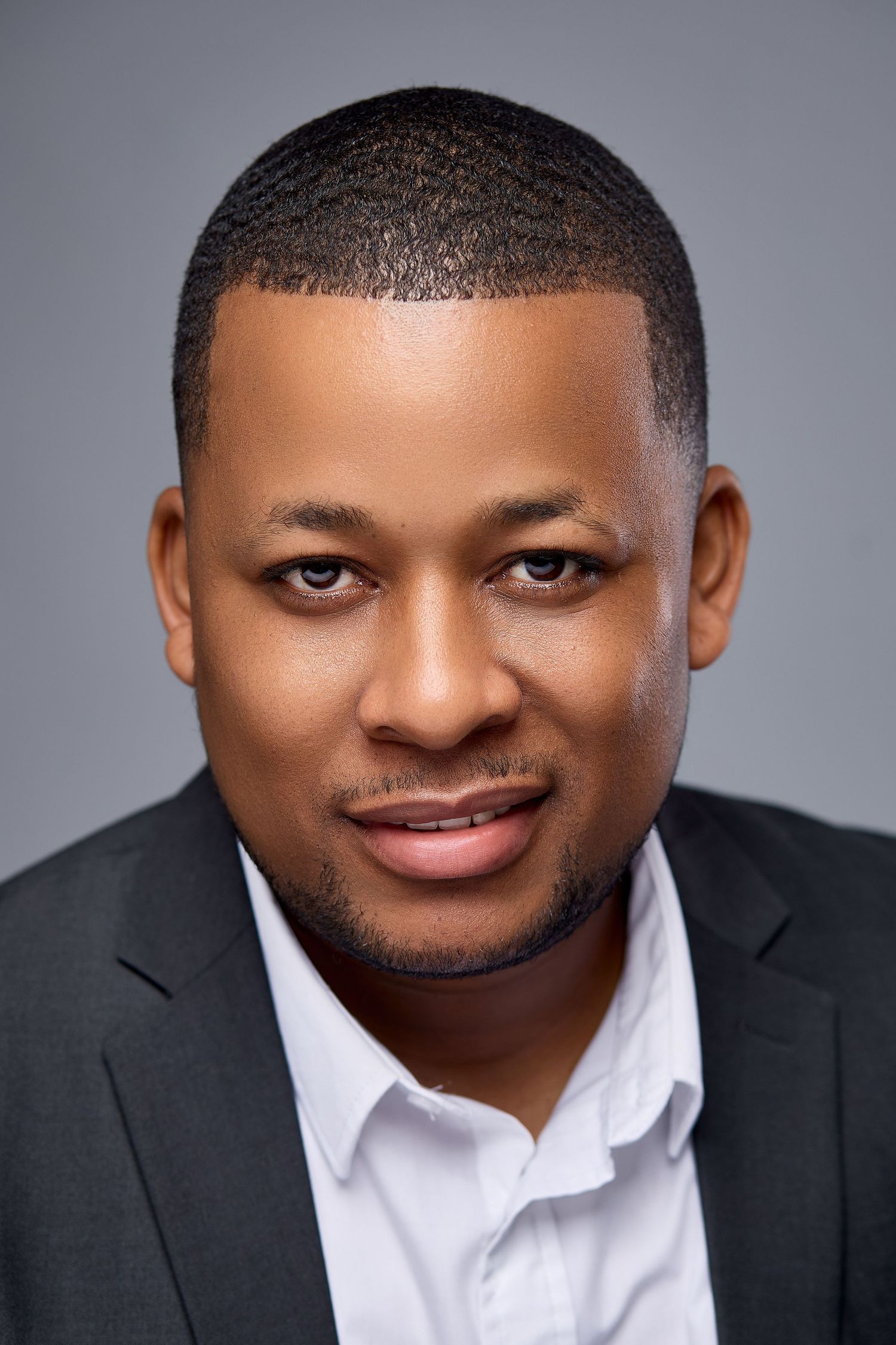 Professional headshot of a smiling young Black man in a dark blazer and white shirt against a gray background.