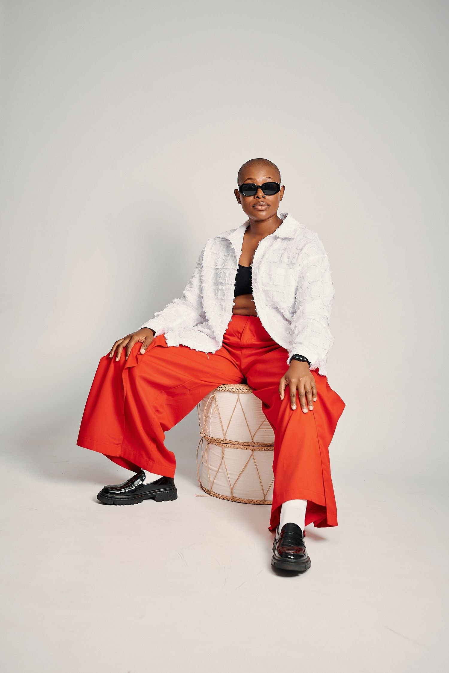Fashionable bald Black woman in white fur jacket and red pants sits on stool wearing sunglasses.