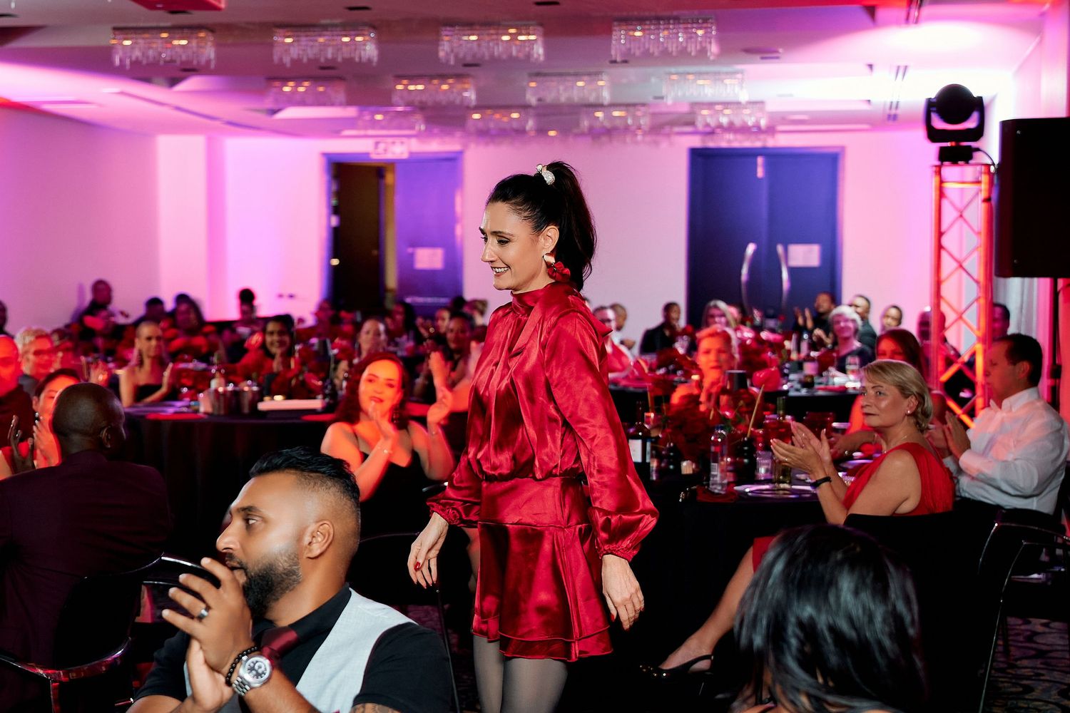 Woman in red satin mini dress walks runway at elegant pink-lit gala event with seated guests.