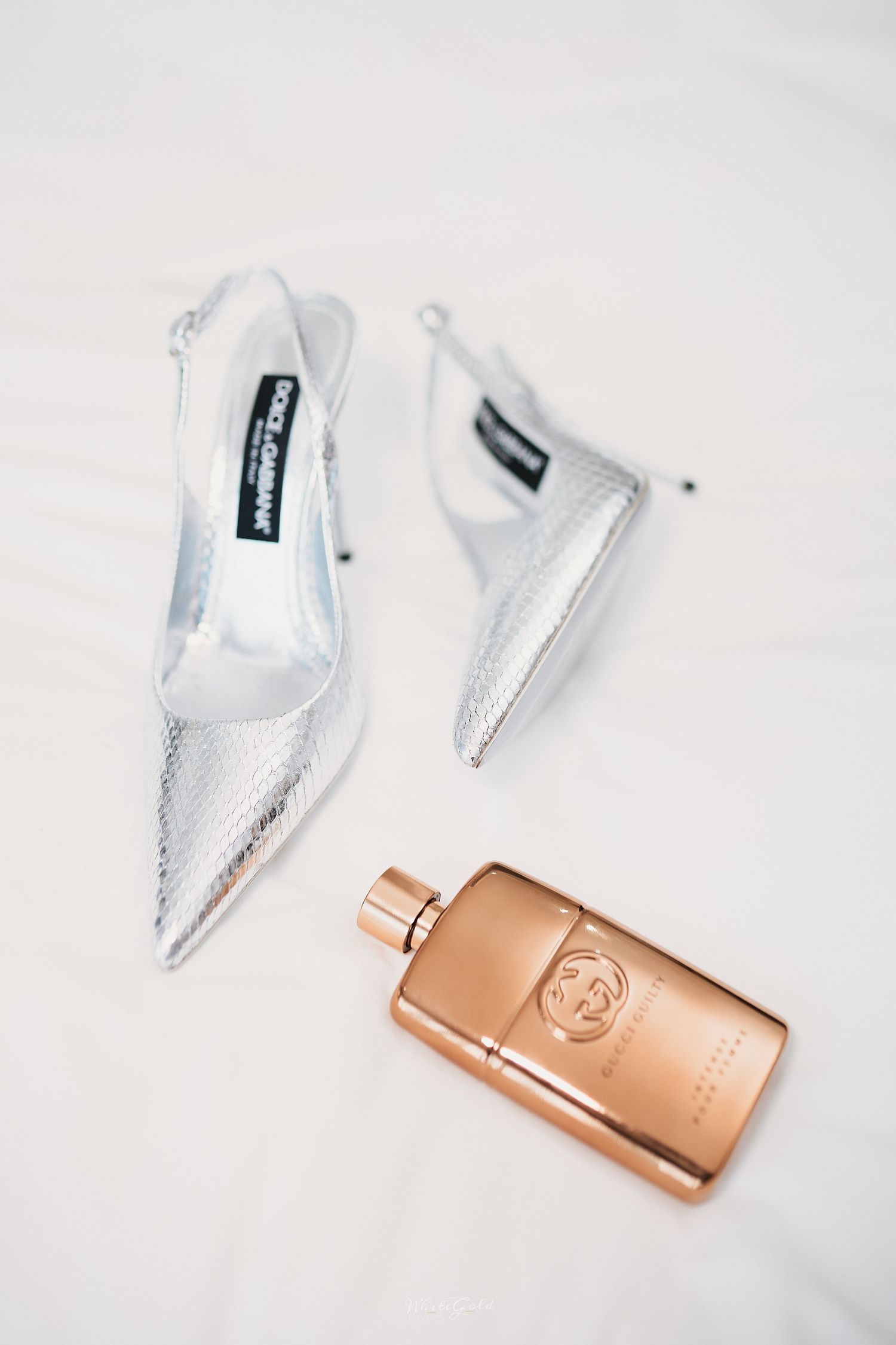 Silver Dolce and Gabbana pointed heels beside a rose gold Gucci perfume bottle on white surface.
