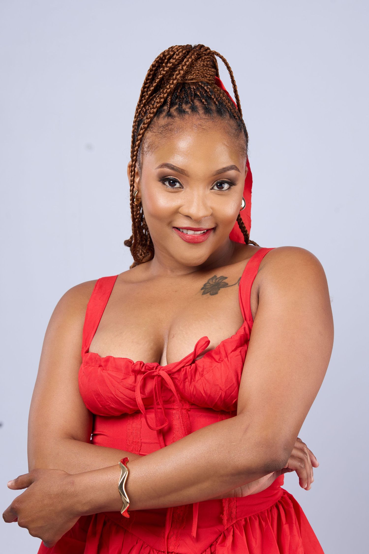 Woman in red dress with braided updo, red earrings, and tattoo, smiling with arms crossed.