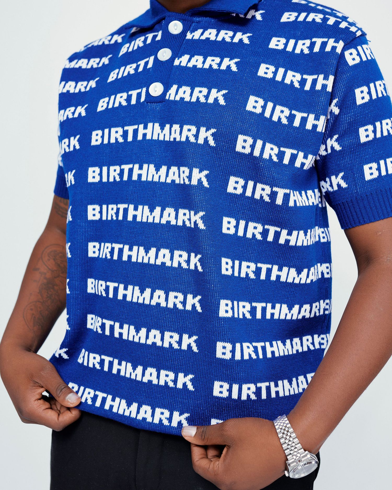 Close-up of blue Birthmark branded knit polo shirt with repeating white logo pattern and silver watch.