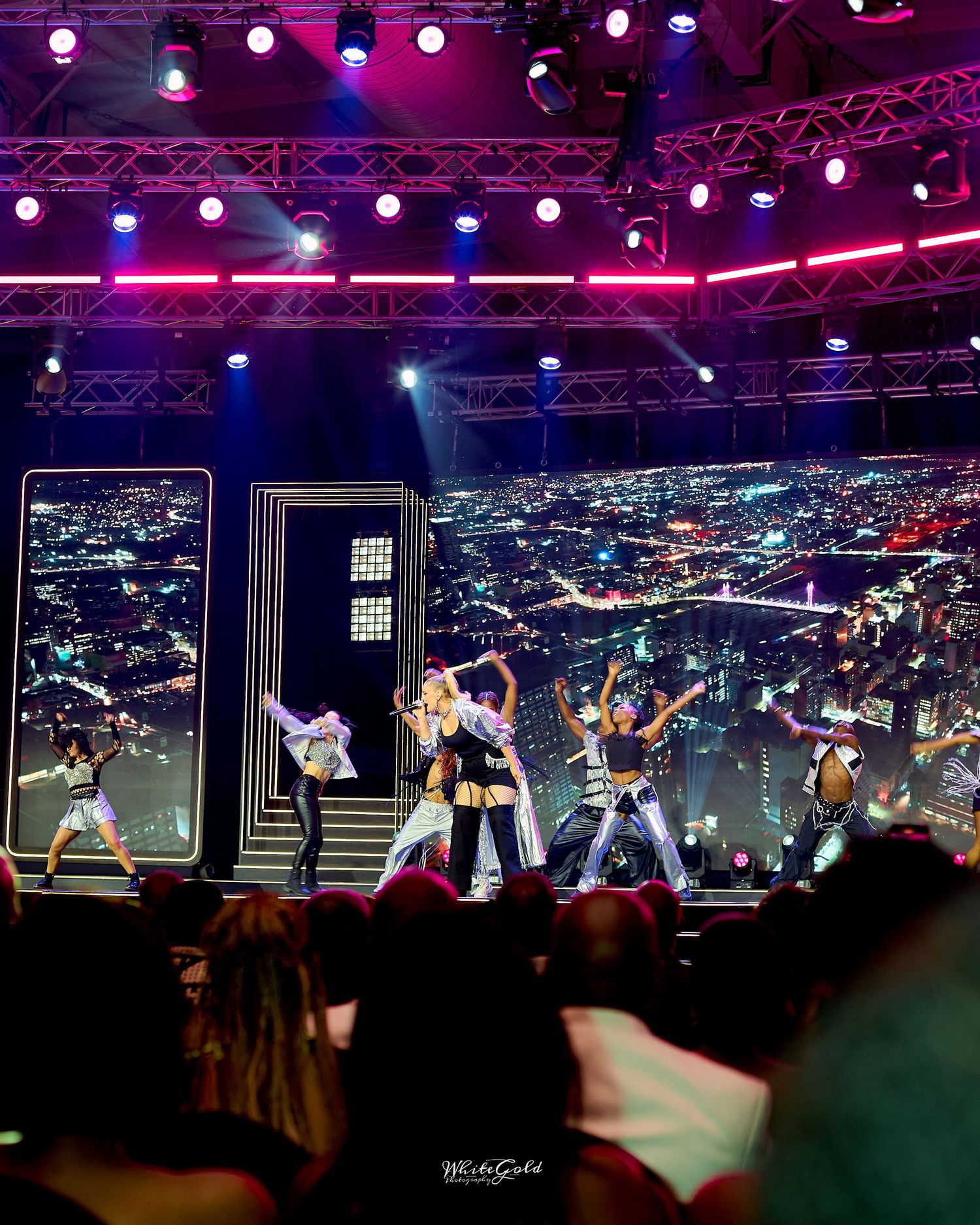 Energetic K-pop style performers on a vibrant stage with city skyline backdrop and colorful lighting.