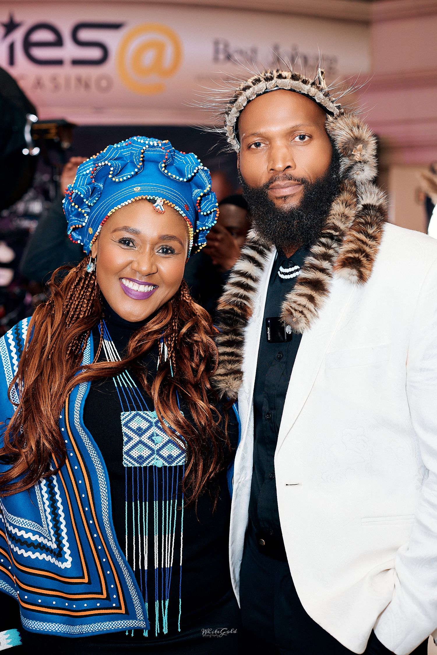 Couple in traditional African attire pose at casino event, woman in blue Ndebele dress and man in white jacket with fur collar.