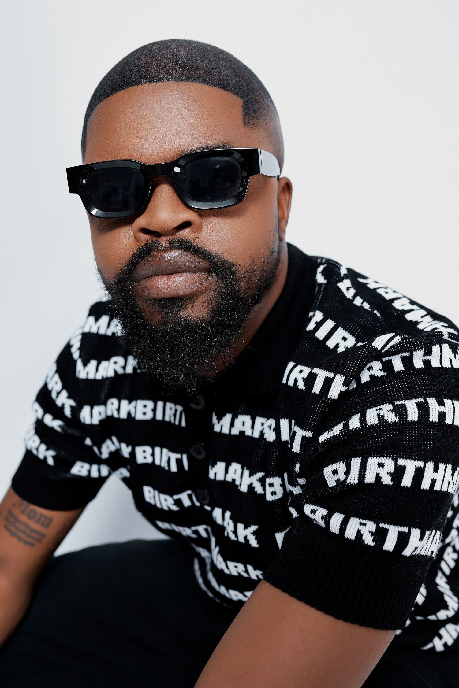 Bearded man wearing black and white Birthmark logo print shirt with dark sunglasses posing.