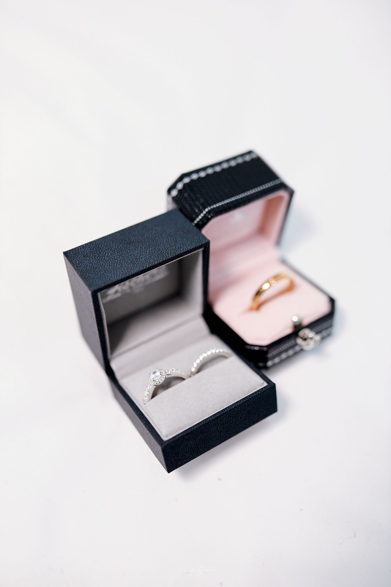 Two open black ring boxes displaying diamond engagement ring and gold wedding band on white surface.