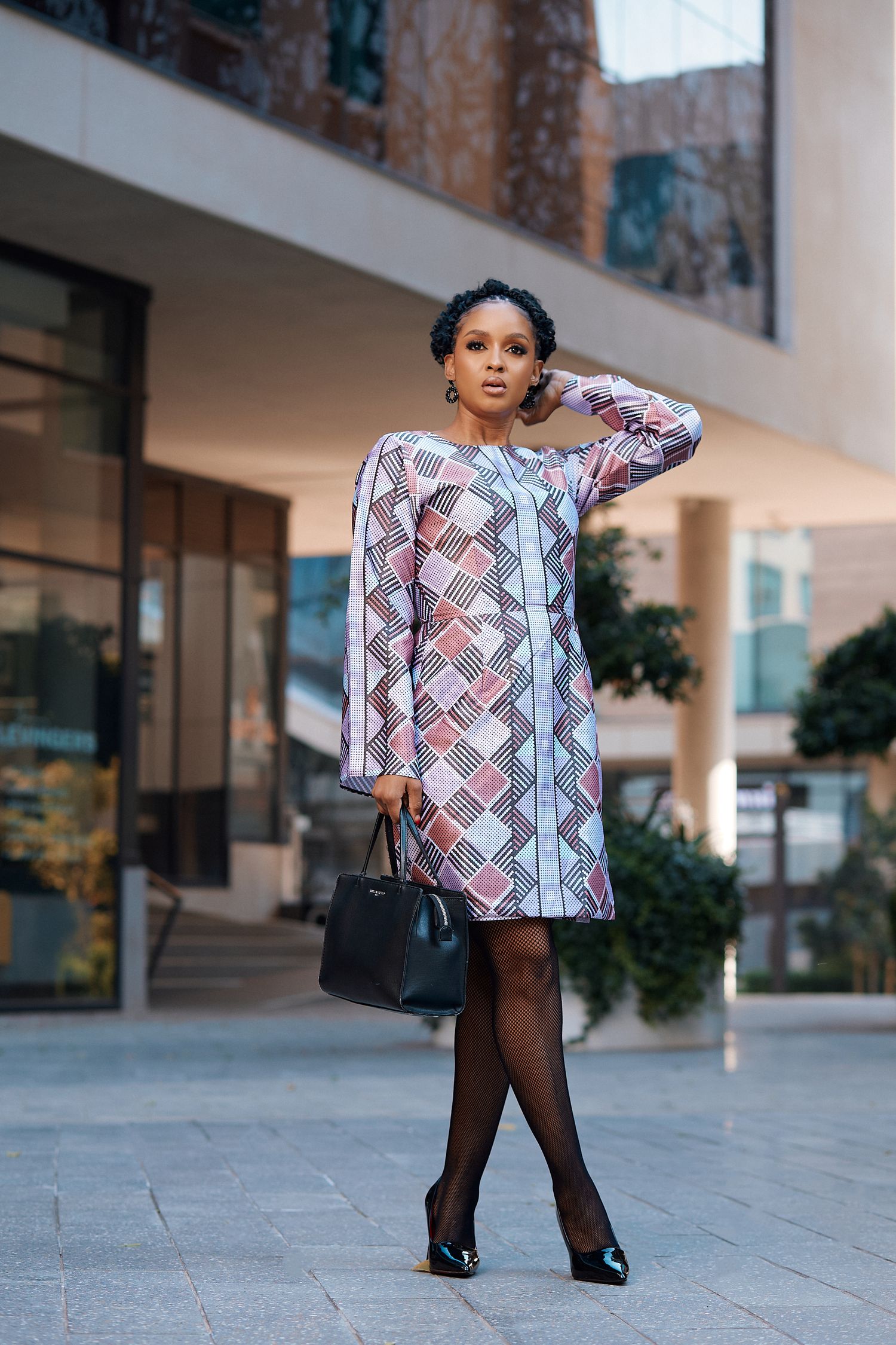 Woman in geometric print shift dress with black tights and heels holding handbag outdoors.