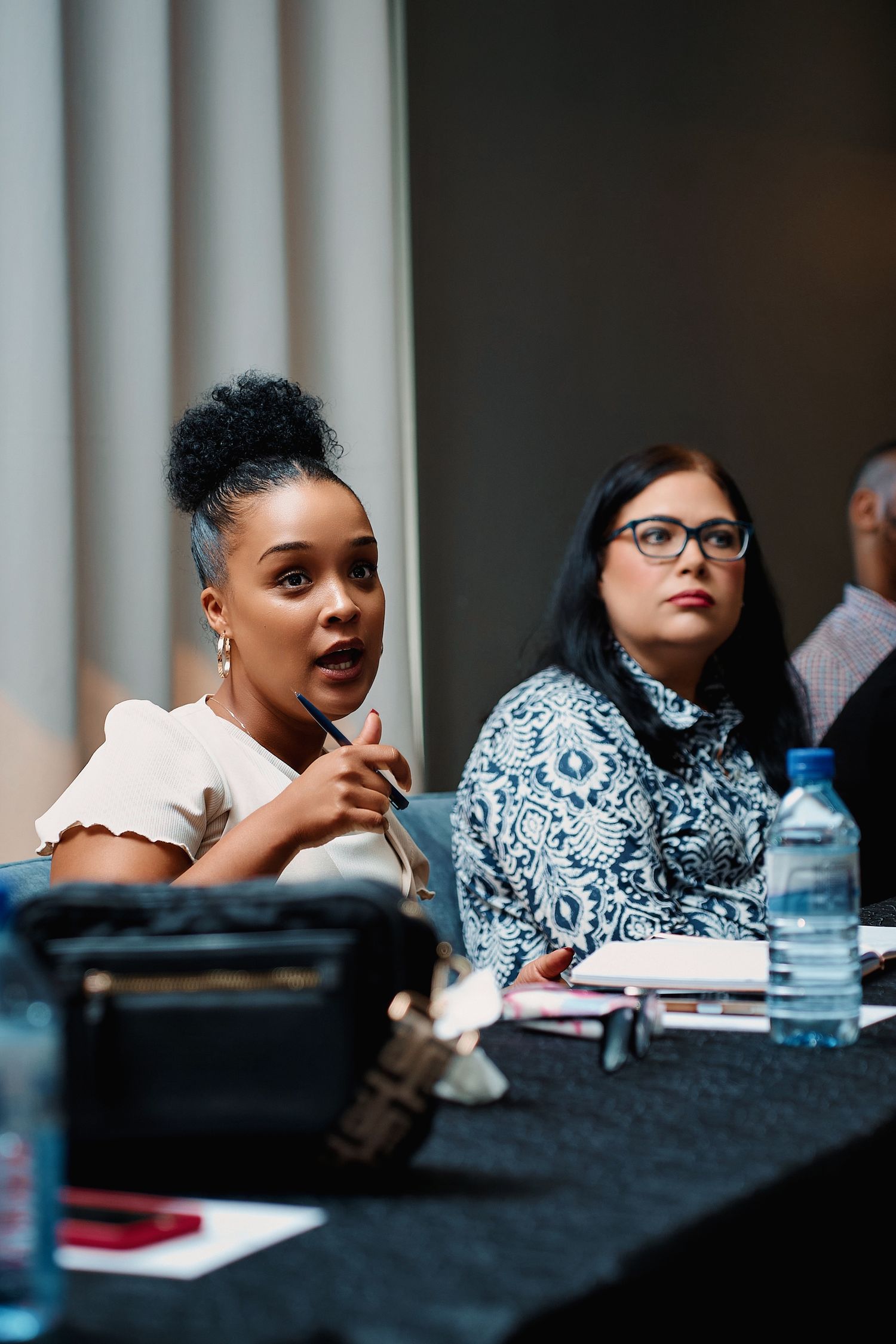 Young Black woman speaking at a panel discussion seated beside a woman in glasses at a conference table.