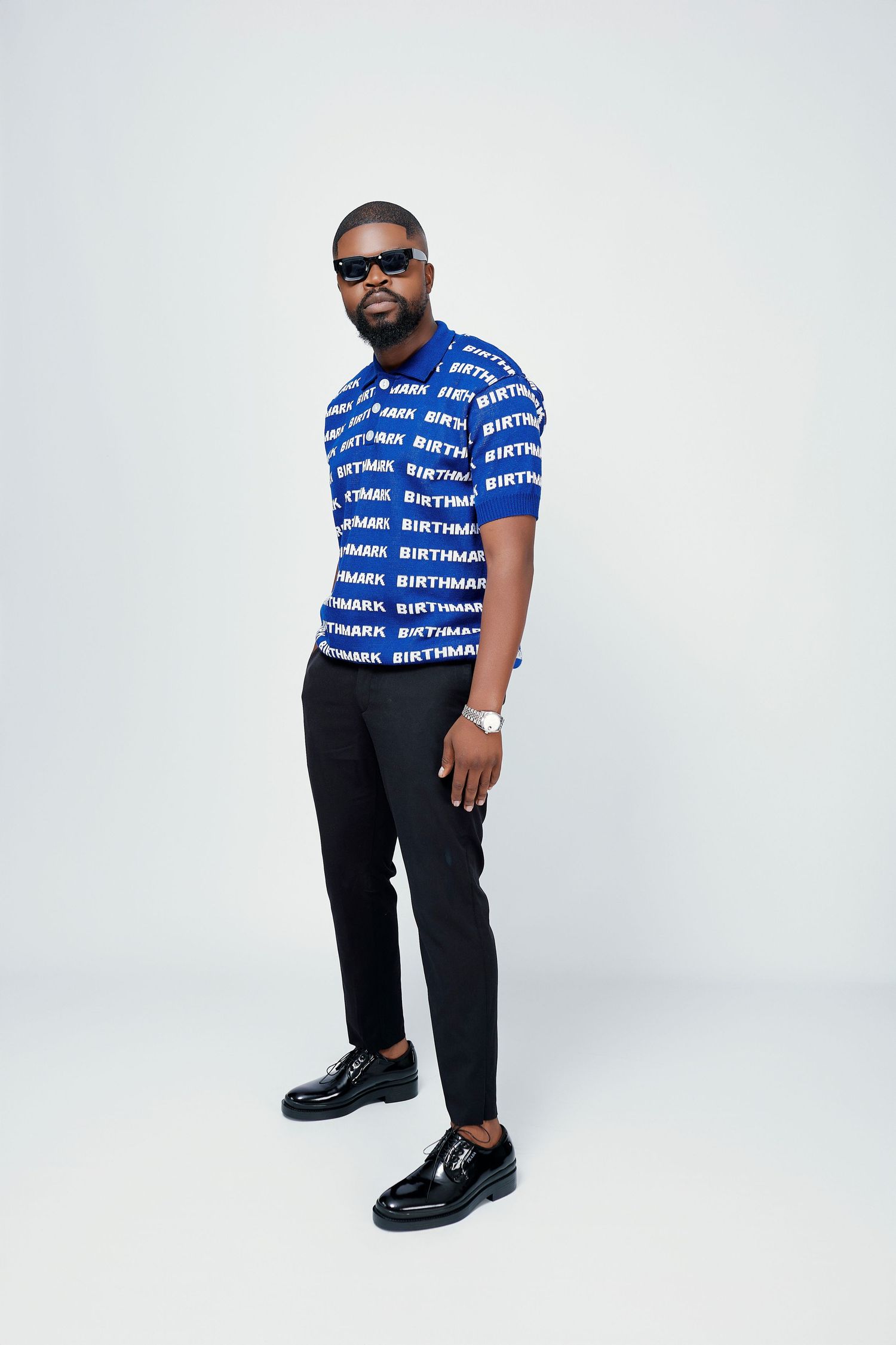Man wearing blue Birthmark logo print polo shirt with black trousers and sunglasses on white background.